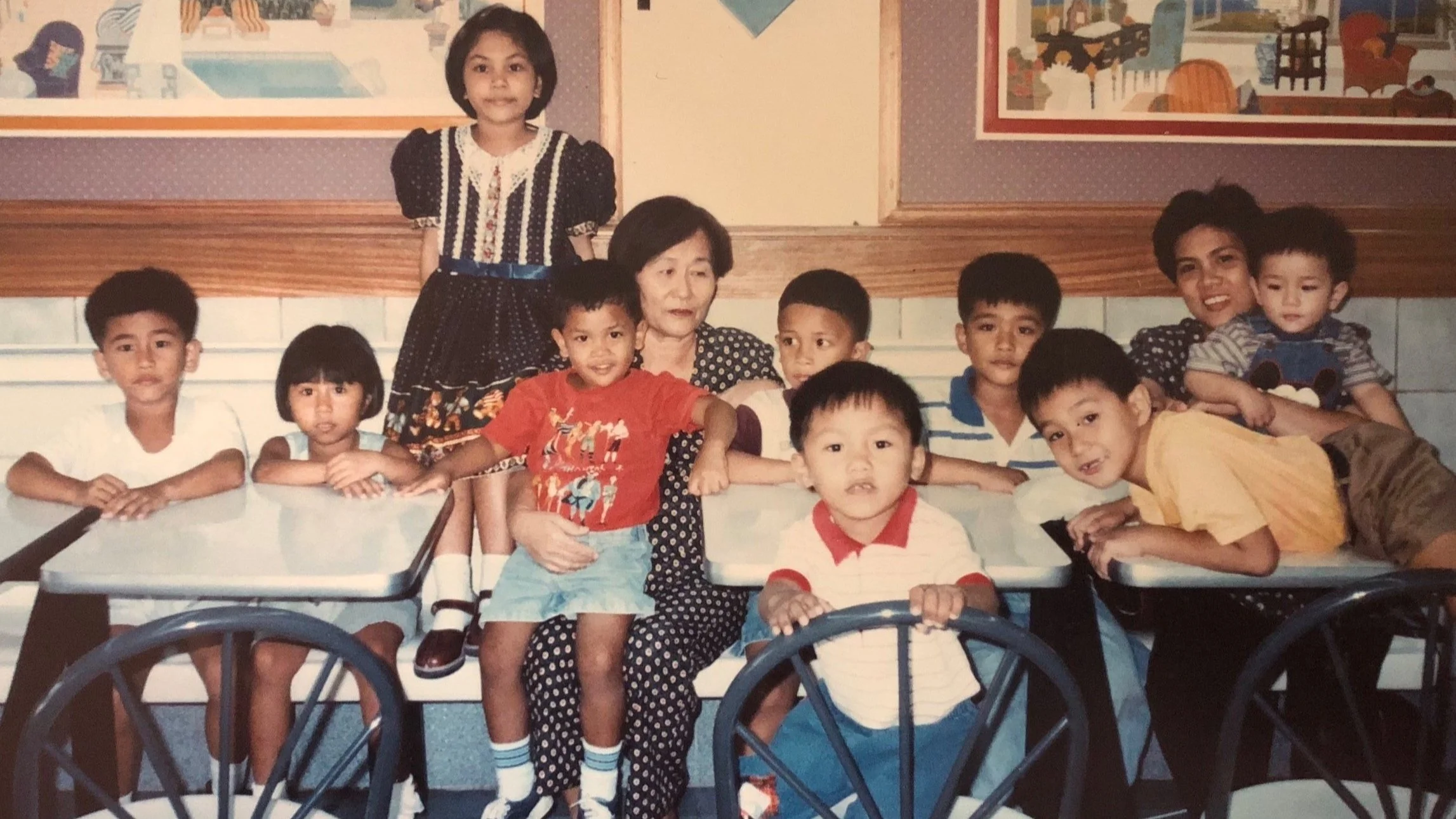 Photo of a young Christopher Reyes sitting on his grandmother's lap. Around them, his cousins are crowded around them. They all smile at the camera.