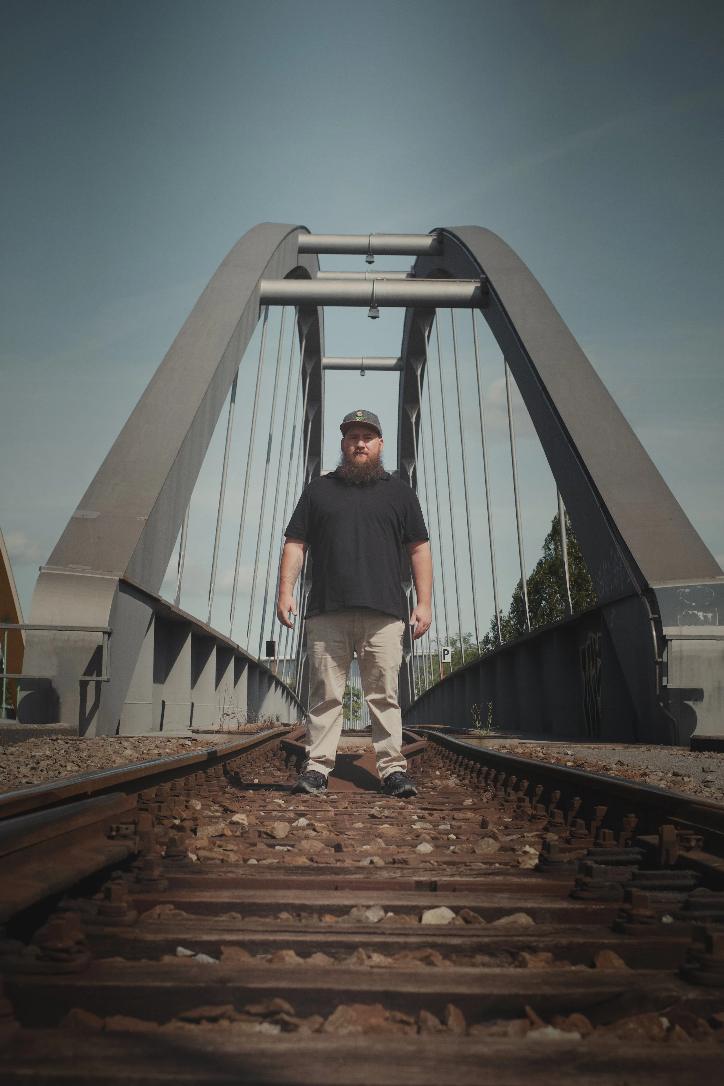 Ein Mann mit Beanie-Hut und Bart steht auf Eisenbahnschienen vor einer modernen Brücke an einem klaren Tag.