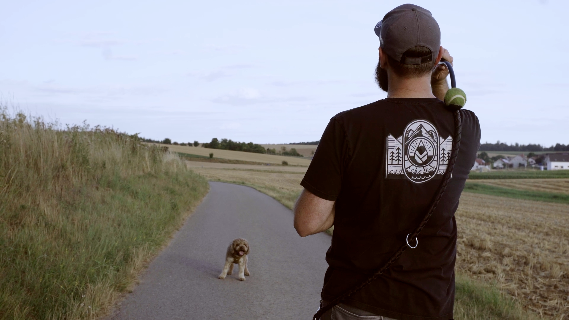 Ein Mann läuft auf einem landwirtschaftlichen Weg, mit einem Hund im Vordergrund. Der Mann trägt ein dunkles T-Shirt mit einem bergigen und naturbezogenen Logo auf dem Rücken.