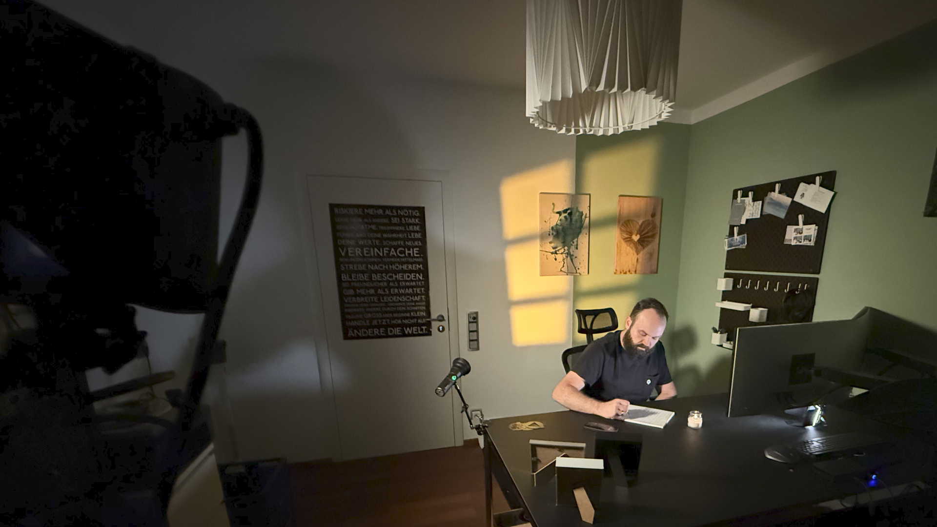 Ein Mann sitzt an einem Tisch in einem Raum mit grüner Wand, einem weißen Lampenschirm und Wanddekorationen. Es gibt einen Computerbildschirm, ein Mikrofon und Kerzen auf dem Tisch.