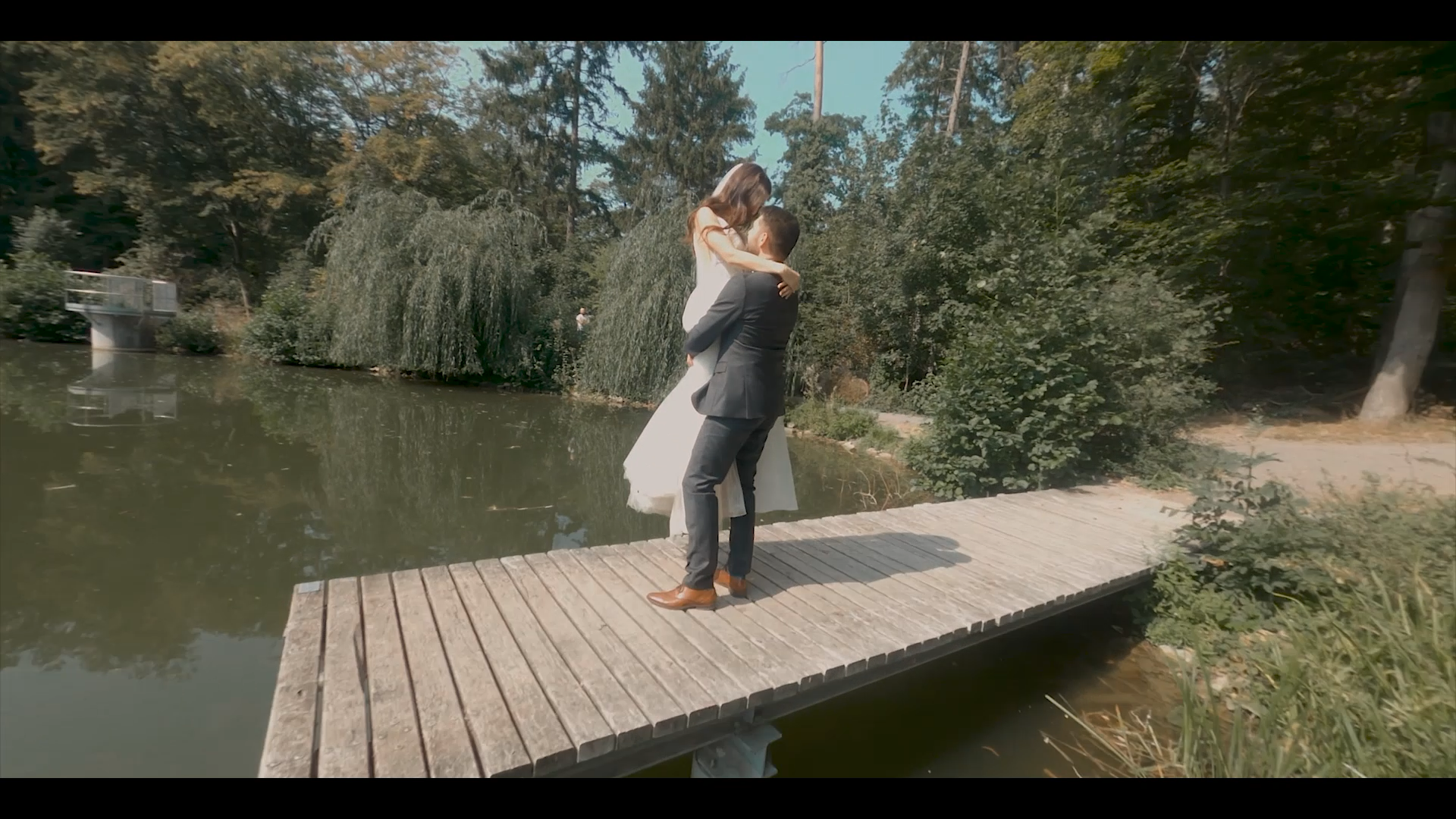 Ein Mann in einem dunklen Anzug hält eine Frau in einem weißen Kleid auf einer Holzbrücke am Wasser hoch, umarmt sie absichtsvoll und schaut sie liebevoll an. Im Hintergrund sind Bäume, strahlend blauer Himmel und Wasser zu sehen.