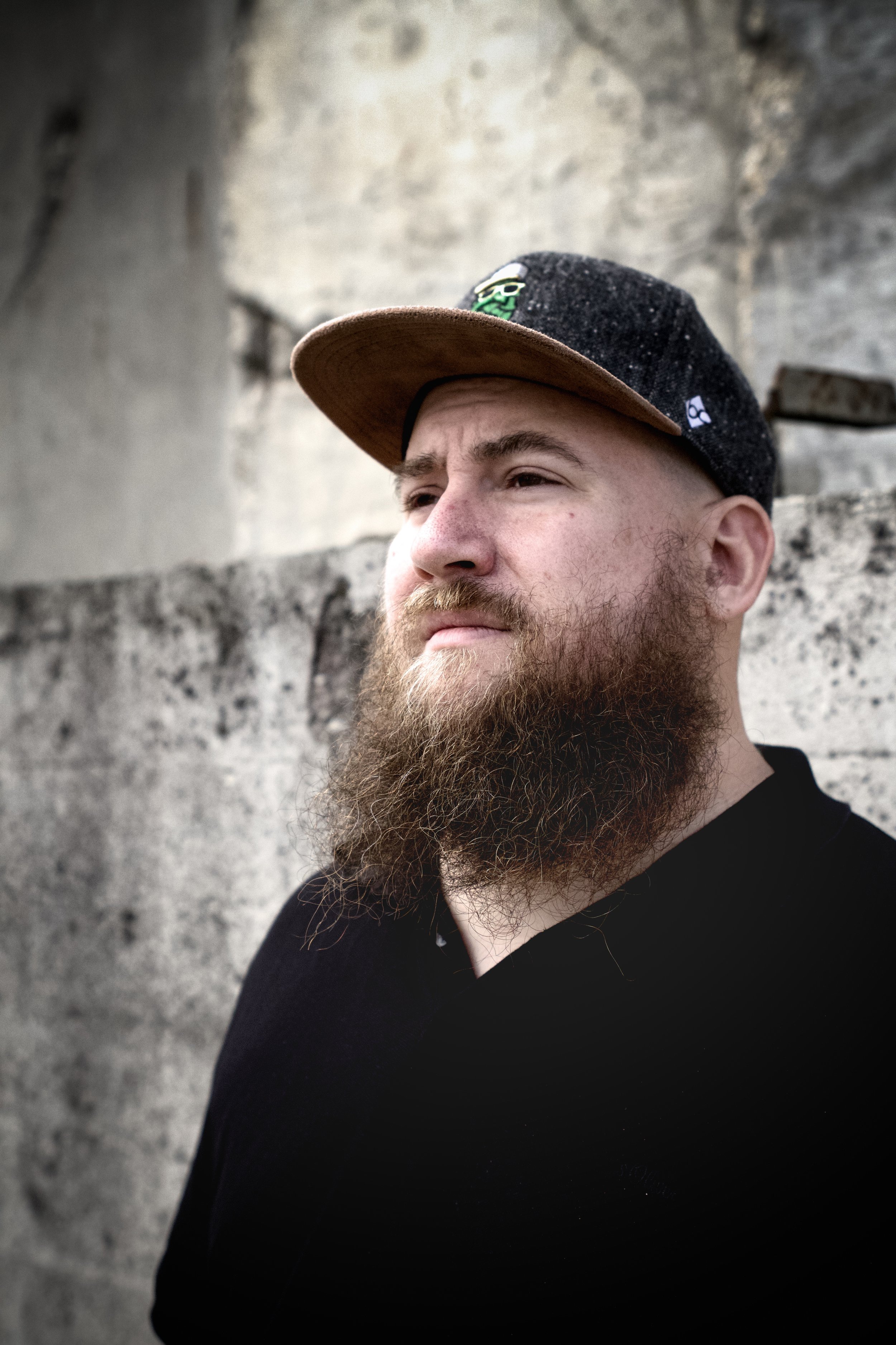 Ein jungen Mann mit Bart trägt eine schwarze Jacke und einen schwarzen Baseballcap mit Filz- und Stoffmaterial, die mit einem grünen und weißen Aufnäher versehen ist, vor einer grauen Steinmauer.