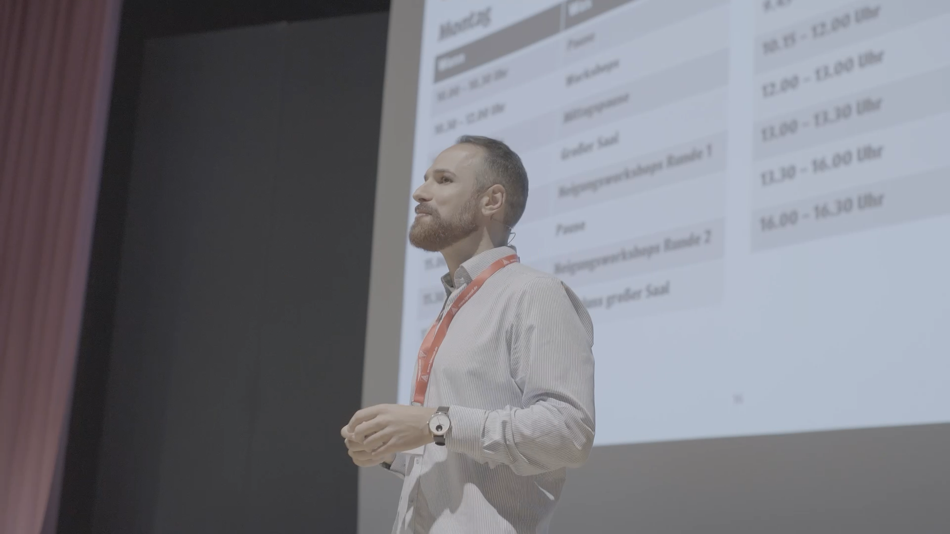 Mann hält einen Vortrag vor einer Präsentationsfolie mit einem Stundenplan. Er trägt ein Hemd, eine Armbanduhr und eine Lanyard. Der Raum ist dunkel mit einem schwarzen Hintergrund und roten Vorhänge links.