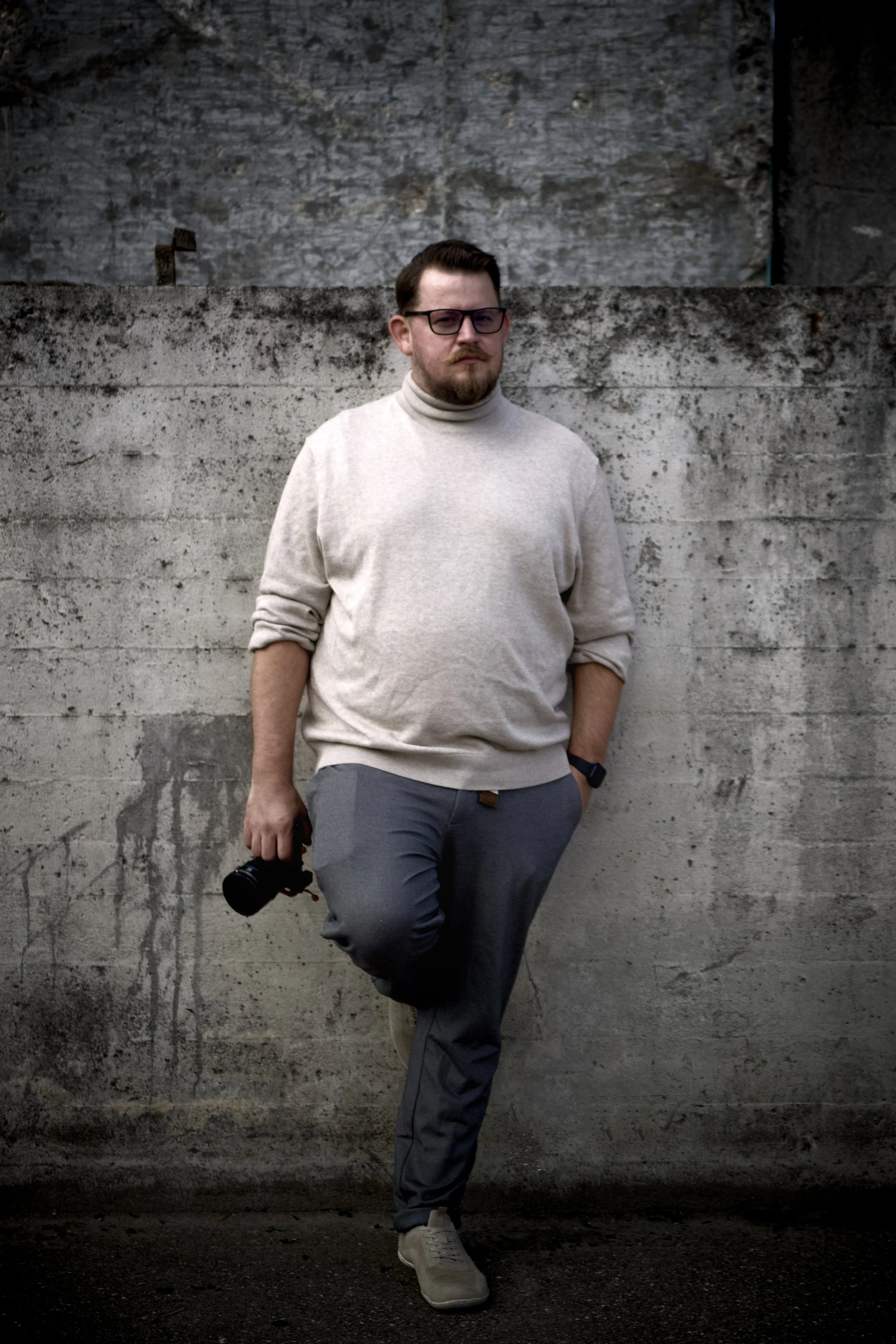 Junger Mann in Brille, Turtleneck-Pullover, grauen Hosen, weißem Schuh, hält eine Kamera, steht vor einer alten, grauen Betonwand.
