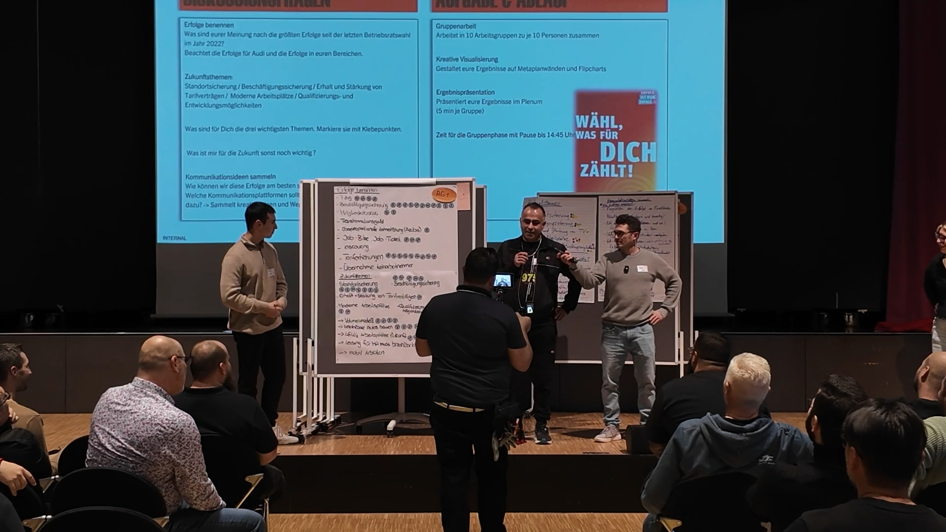 Eine Bühne mit mehreren Personen, die vor einem Publikum sprechen. Im Hintergrund ein großes Bildschirm mit Text und einem Slogan, sowie drei Whiteboards mit Notizen. Das Publikum sitzt im Raum und sieht zu.