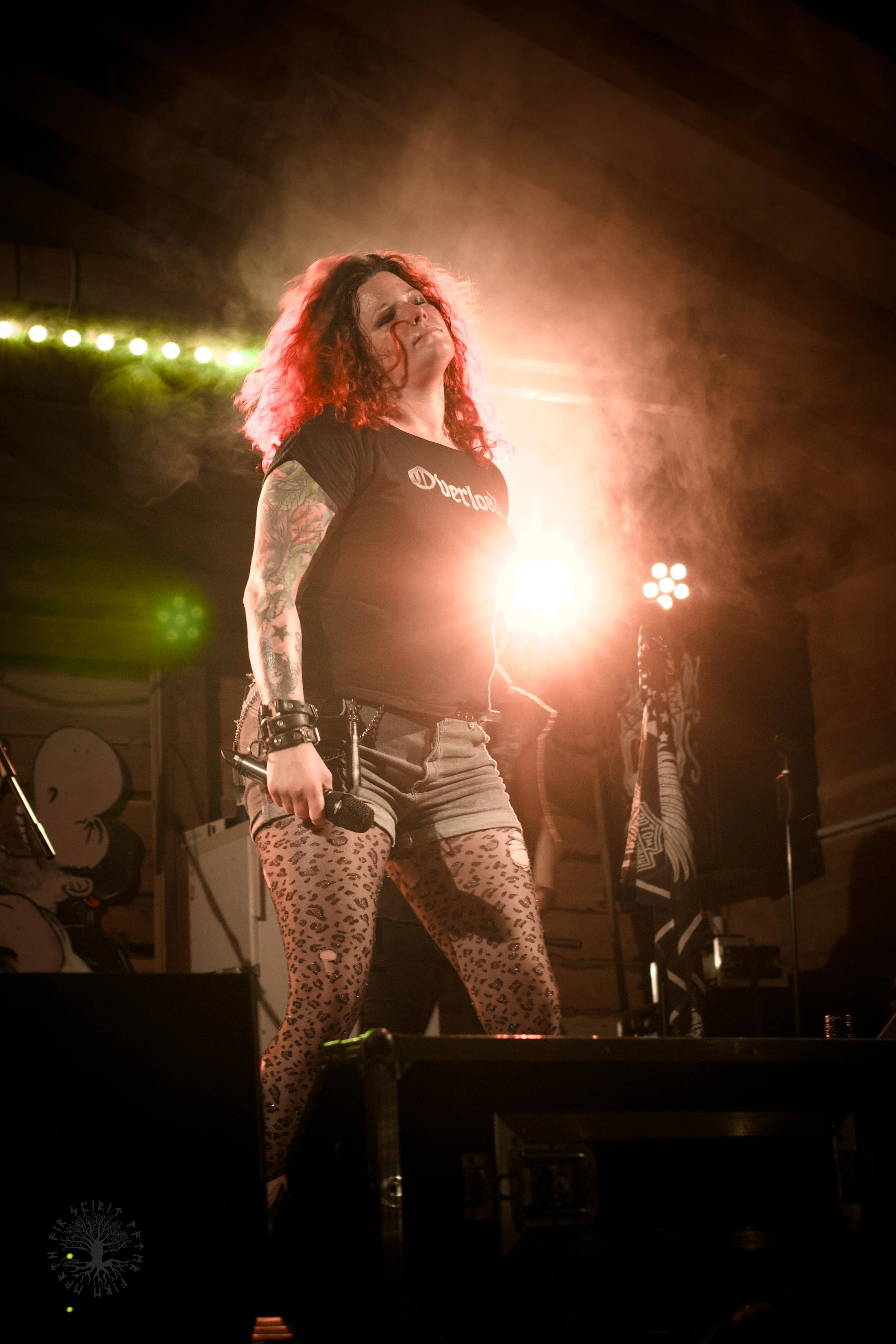 Eine Frau mit lockigem Haar, Tattoos auf den Armen, trägt einen schwarzen T-Shirt mit weißem Logo und Leoparden-leggings, steht auf einer Bühne vor bunten Lichtern, während sie Musik macht.