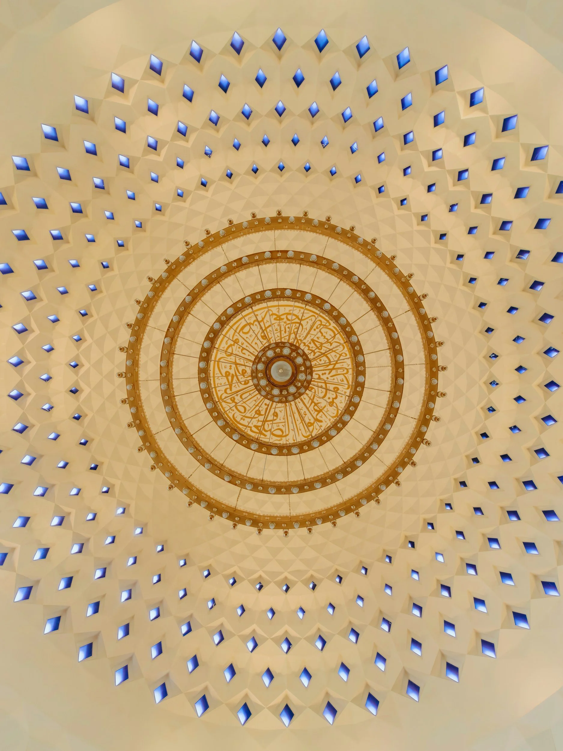 Looking up at a dome ceiling with a geometric pattern of illuminated blue squares and a central circular chandelier with Arabic calligraphy.