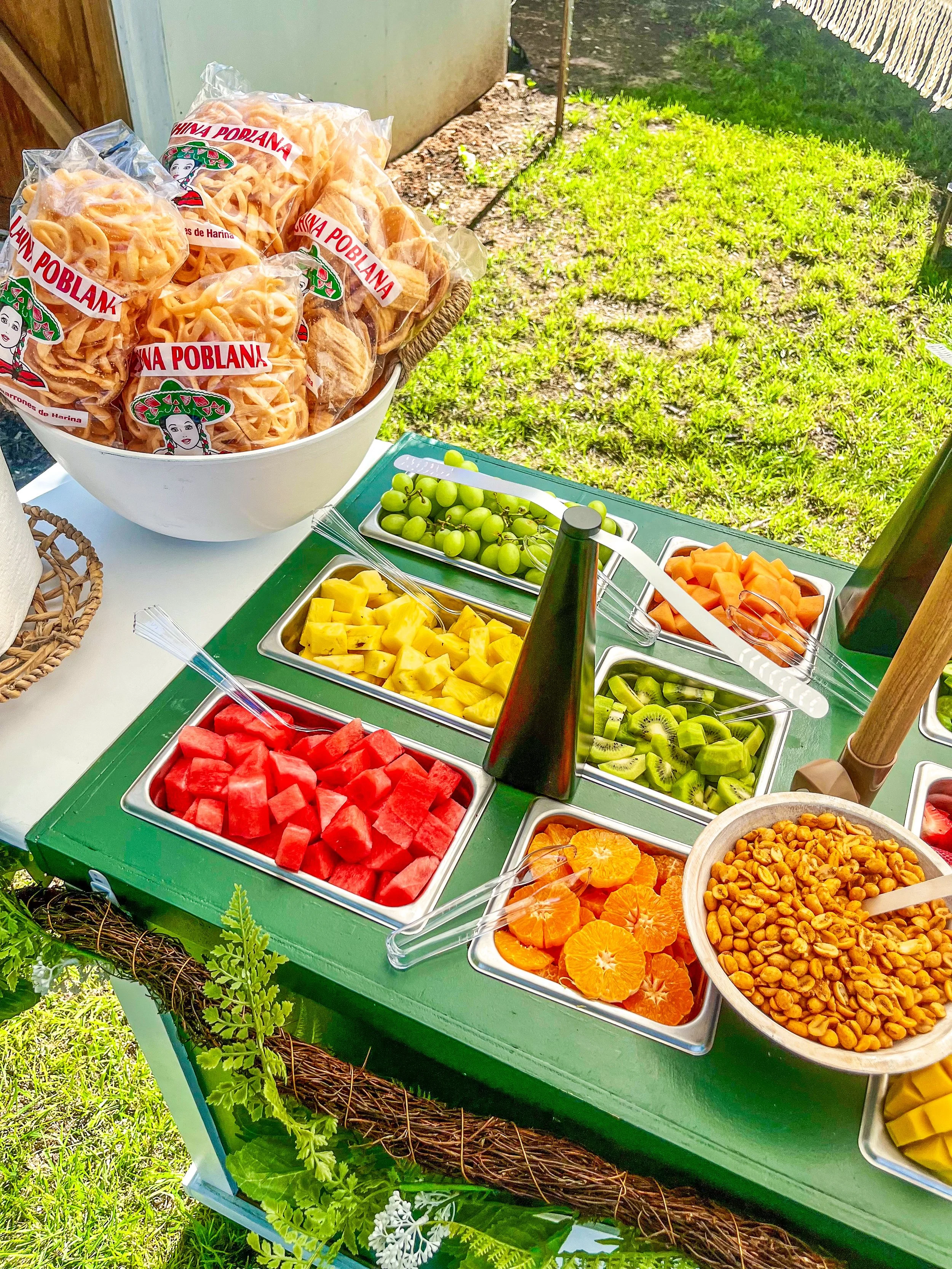 A table filled with bowls of cut fruit including watermelon, pineapple, mandarin oranges, kiwi, cantaloupe, and green grapes, with a large bowl of packaged toasted tortillas labeled 'China Poblana' and a bottle of beverage, set outdoors on grass.