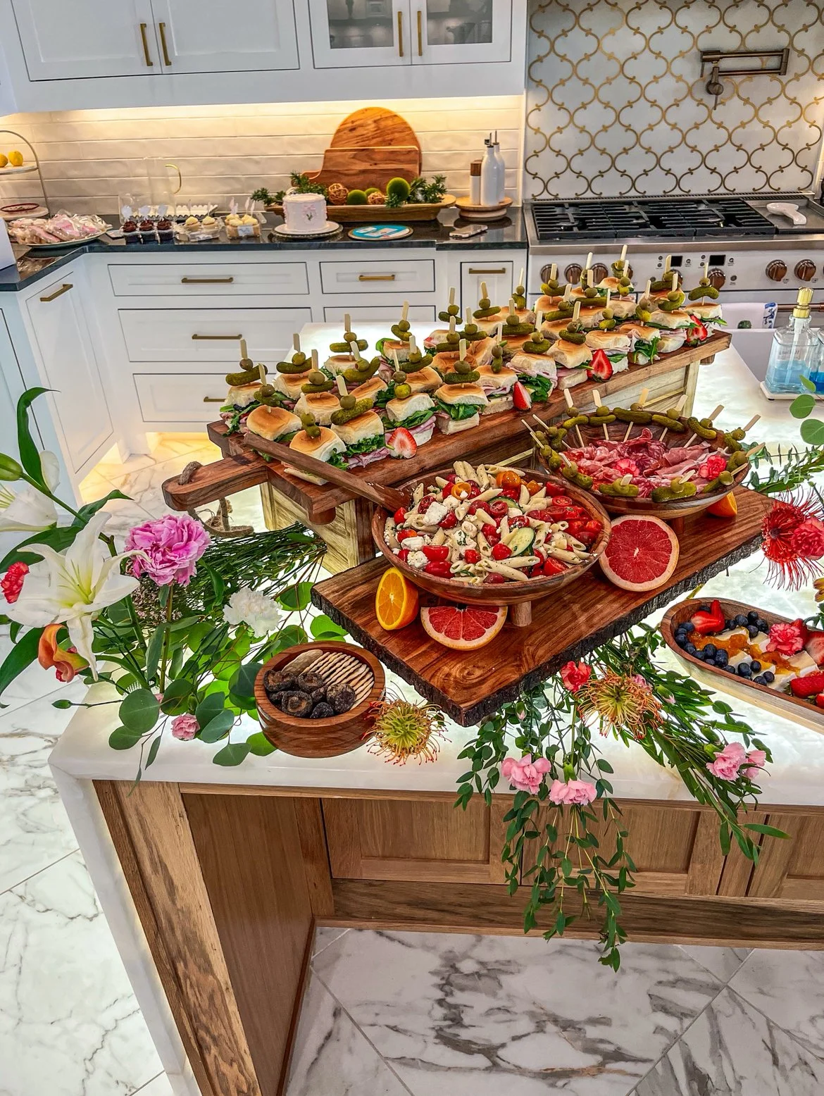 A kitchen island decorated with a floral arrangement and a variety of appetizers, including sandwiches with skewers, pasta salad, and fruit. The background shows a white kitchen with a cake, desserts, and a cutting board on the countertop.