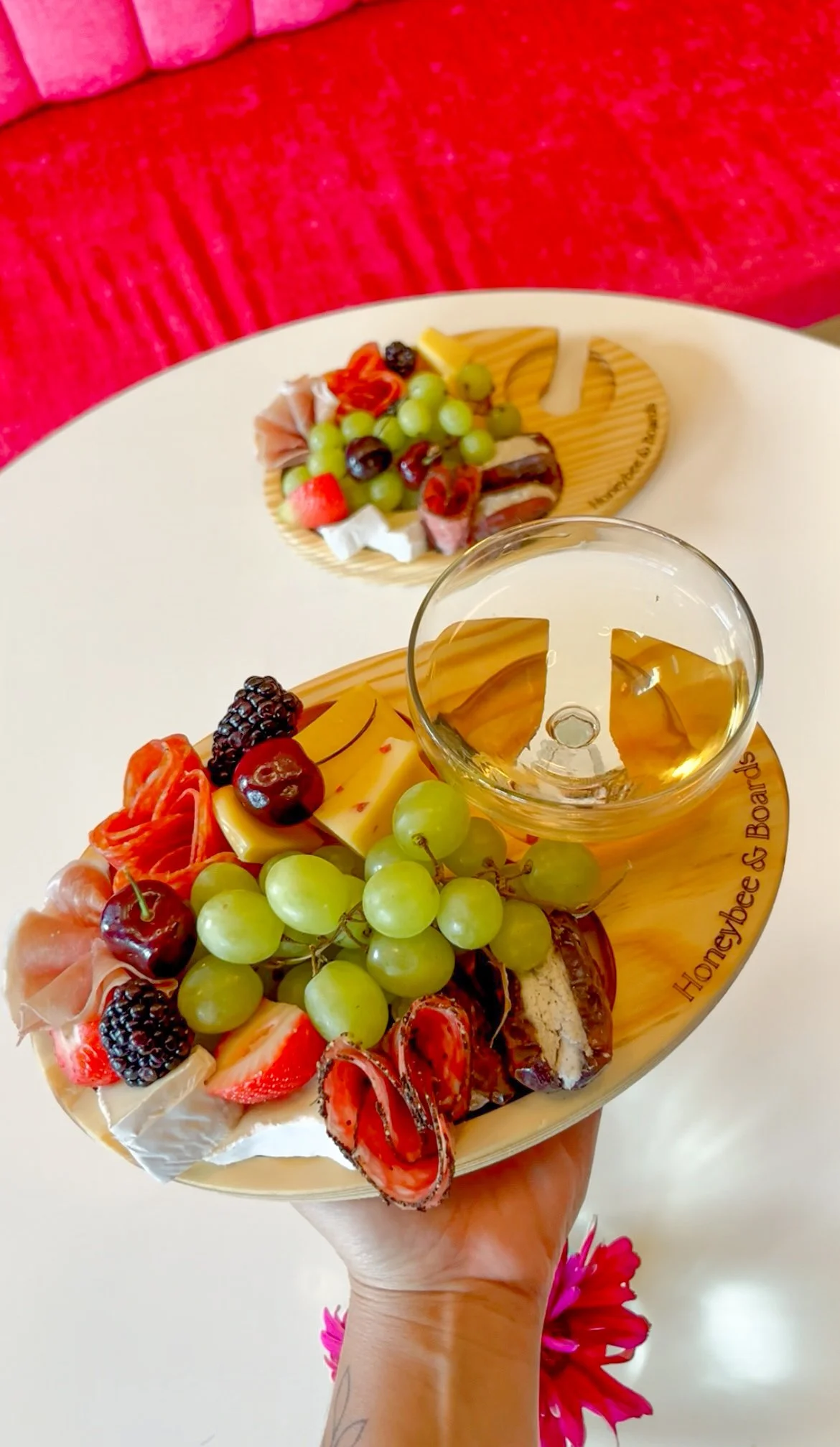 A hand holding a small cheese and fruit plate with green grapes, strawberries, blackberries, cherries, slices of cheese, and meat, with a glass of white wine on a wooden honeycomb shaped serving board. Another similar plate with fruit and cheese is i