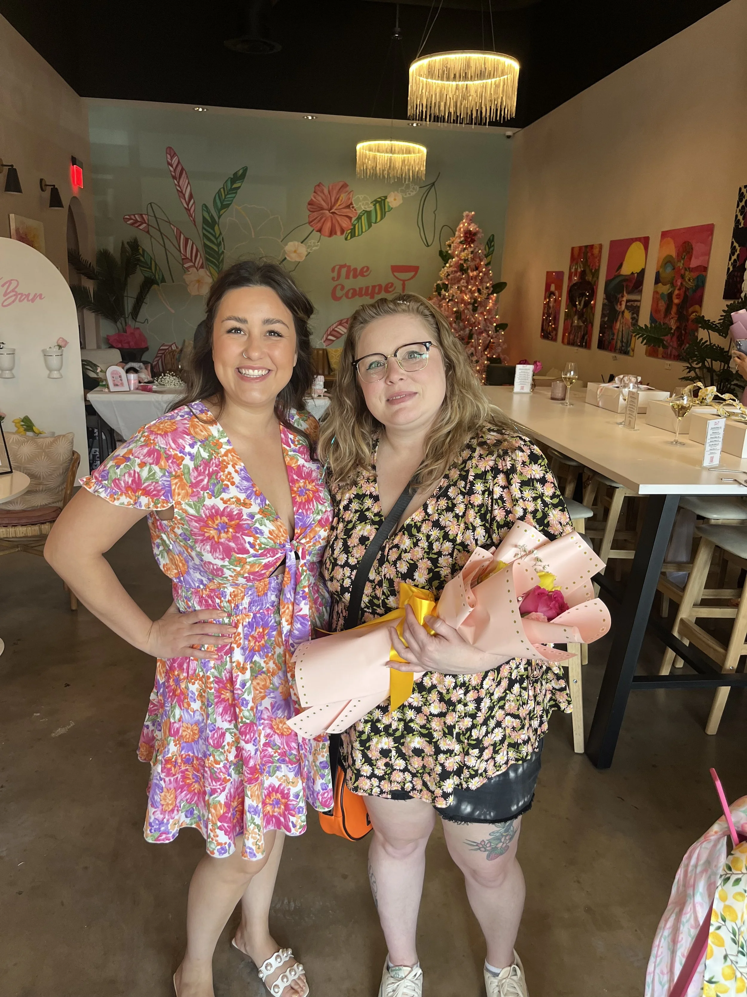 Two women smiling in a decorated indoor setting, one holding a bouquet of flowers.