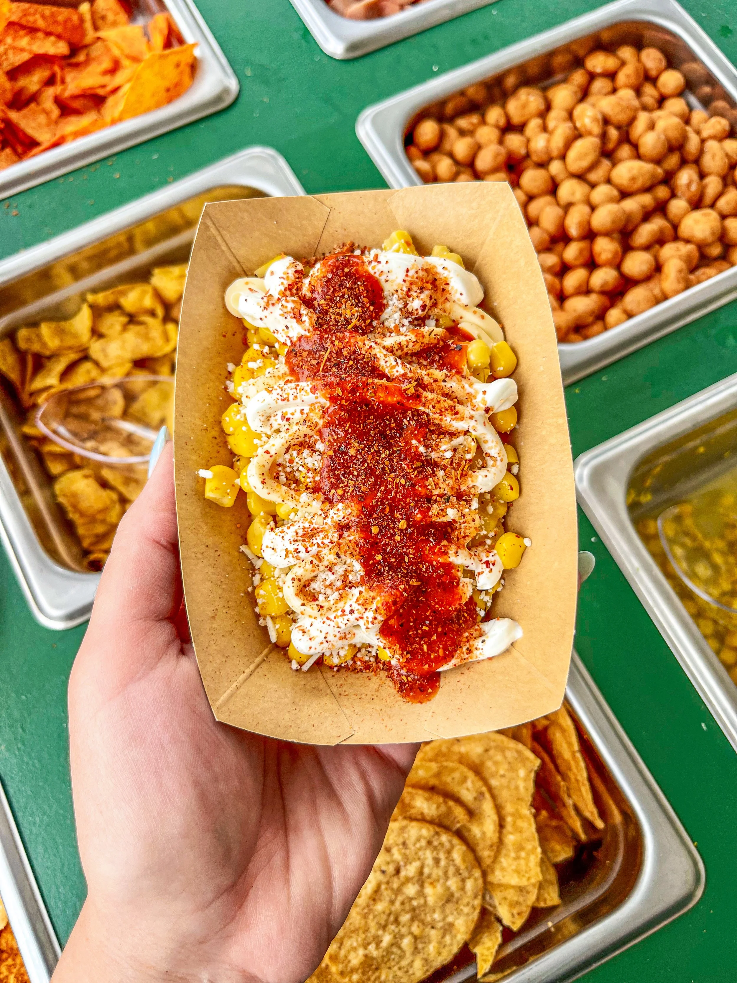 A hand holding a tray of corn topped with mayonnaise, red chili powder, and hot sauce, with various hot foods in serving trays in the background.