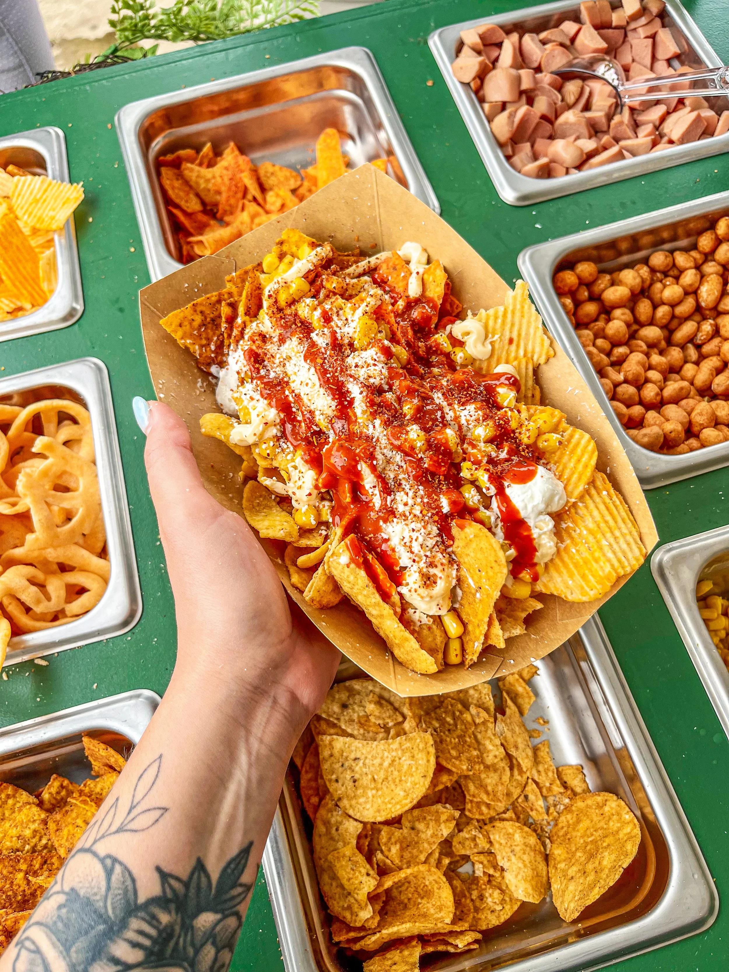 A hand holding a tray of loaded nachos topped with cheese, sour cream, corn, salsa, and hot sauce, in front of a selection of toppings including diced hot dogs, chili, seasoned meat, and other condiments.
