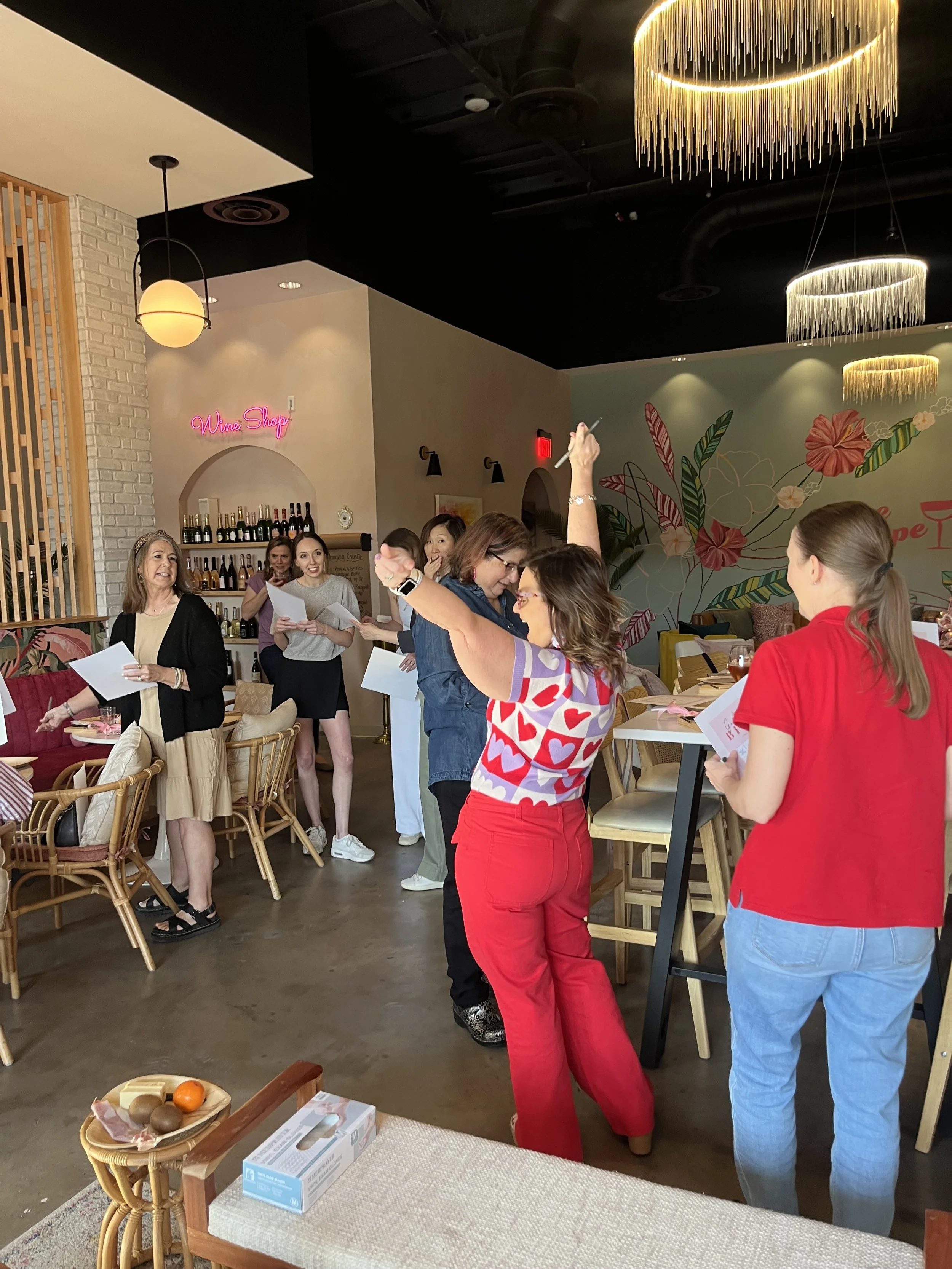 A group of women gathered in a restaurant, some holding papers and engaging with each other, with one woman in red raising her arm. The restaurant has a colorful interior with decorative wall art, a sign that reads 'Wine Shop', and elegant lighting f
