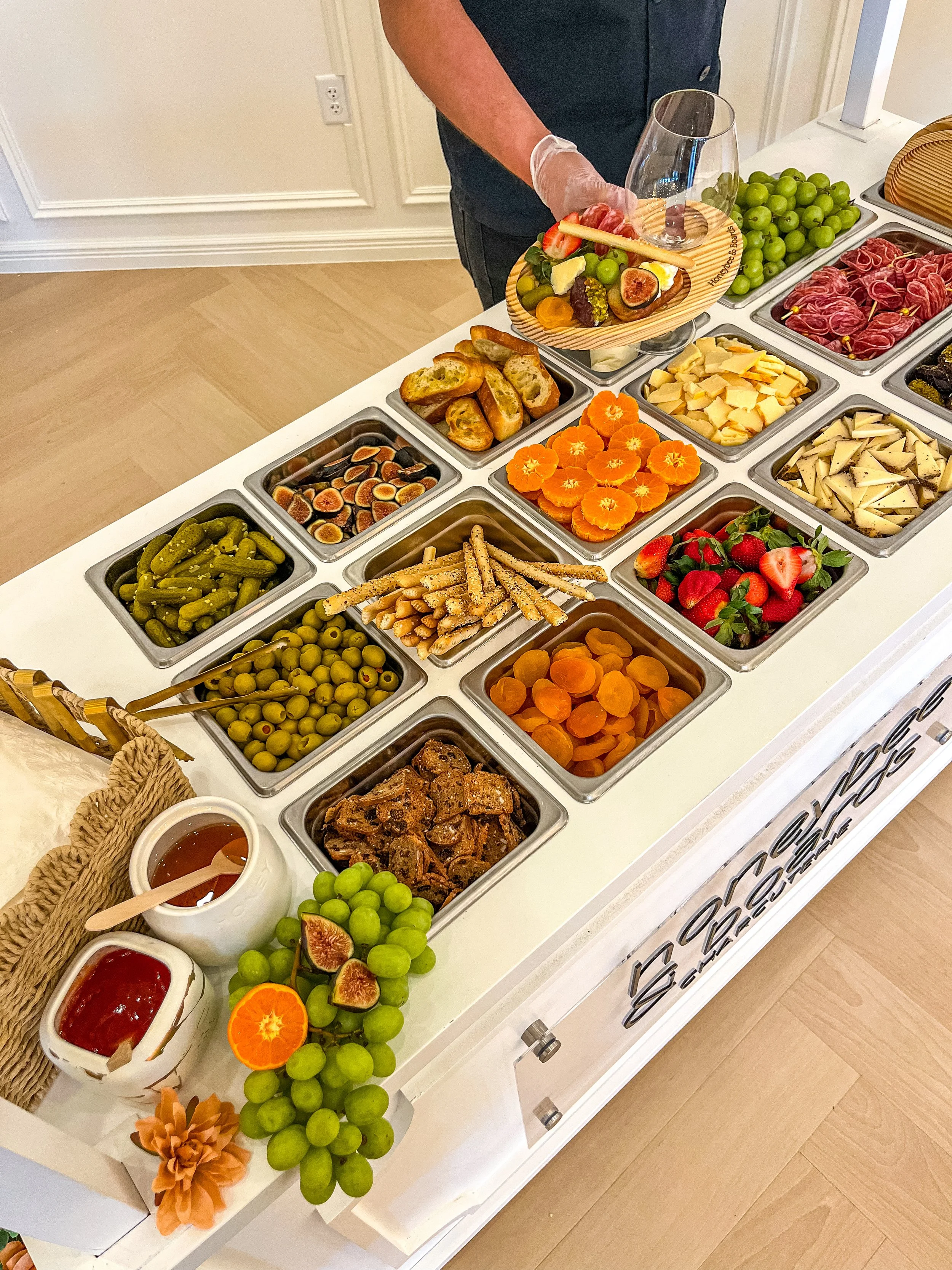 A buffet table with various fruits, cheeses, and snacks, including grapes, strawberries, oranges, figs, sliced cheeses, pickles, and bread, with a person serving themselves.