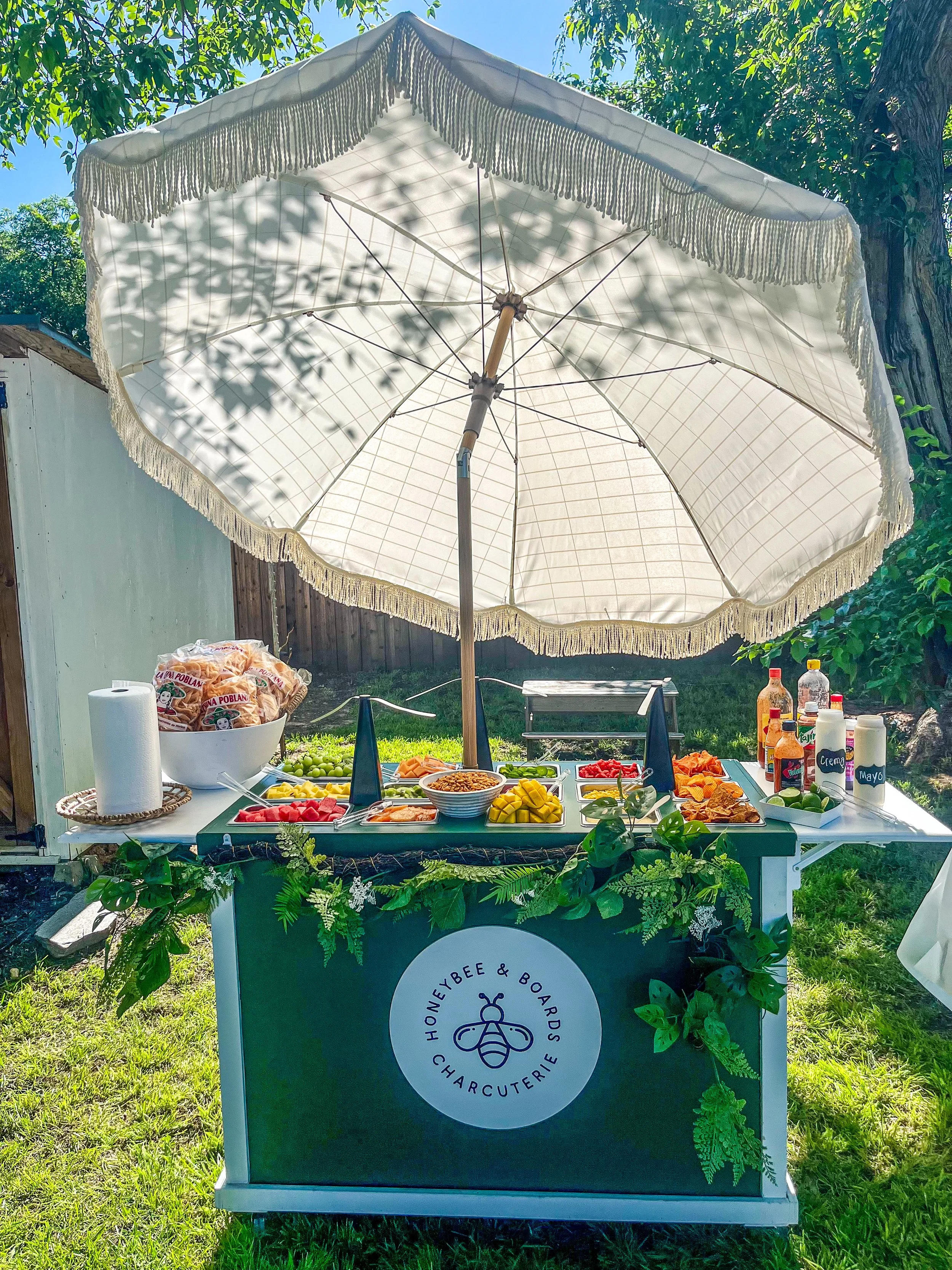 Outdoor food stand with a white umbrella providing shade, offering various fresh fruits, vegetables, and condiments for a barbecue or picnic, decorated with greenery, featuring a logo that reads 'HONEYBEE & BOARDS CHAR CUTERIE' with a bee illustratio
