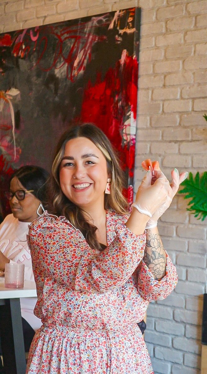 A smiling woman with tattoos on her right arm and earrings, wearing a floral dress, holds a small spoon near her face inside a restaurant with a brick wall and abstract painting in the background. Another woman is seated at the table in the backgroun