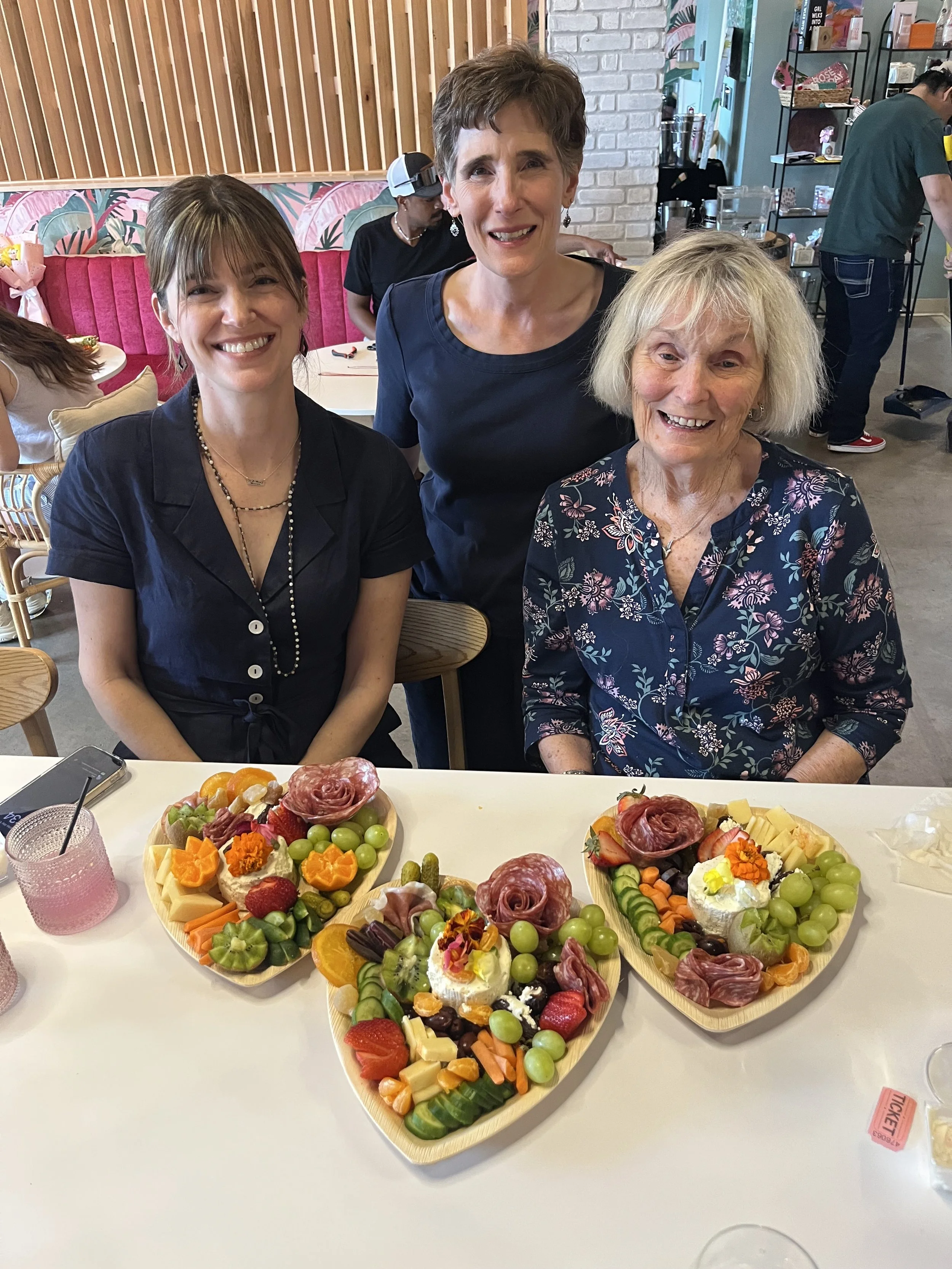 Three women sitting at a table with three heart-shaped platters of assorted cheese, fruits, and meats, smiling, in a casual restaurant.