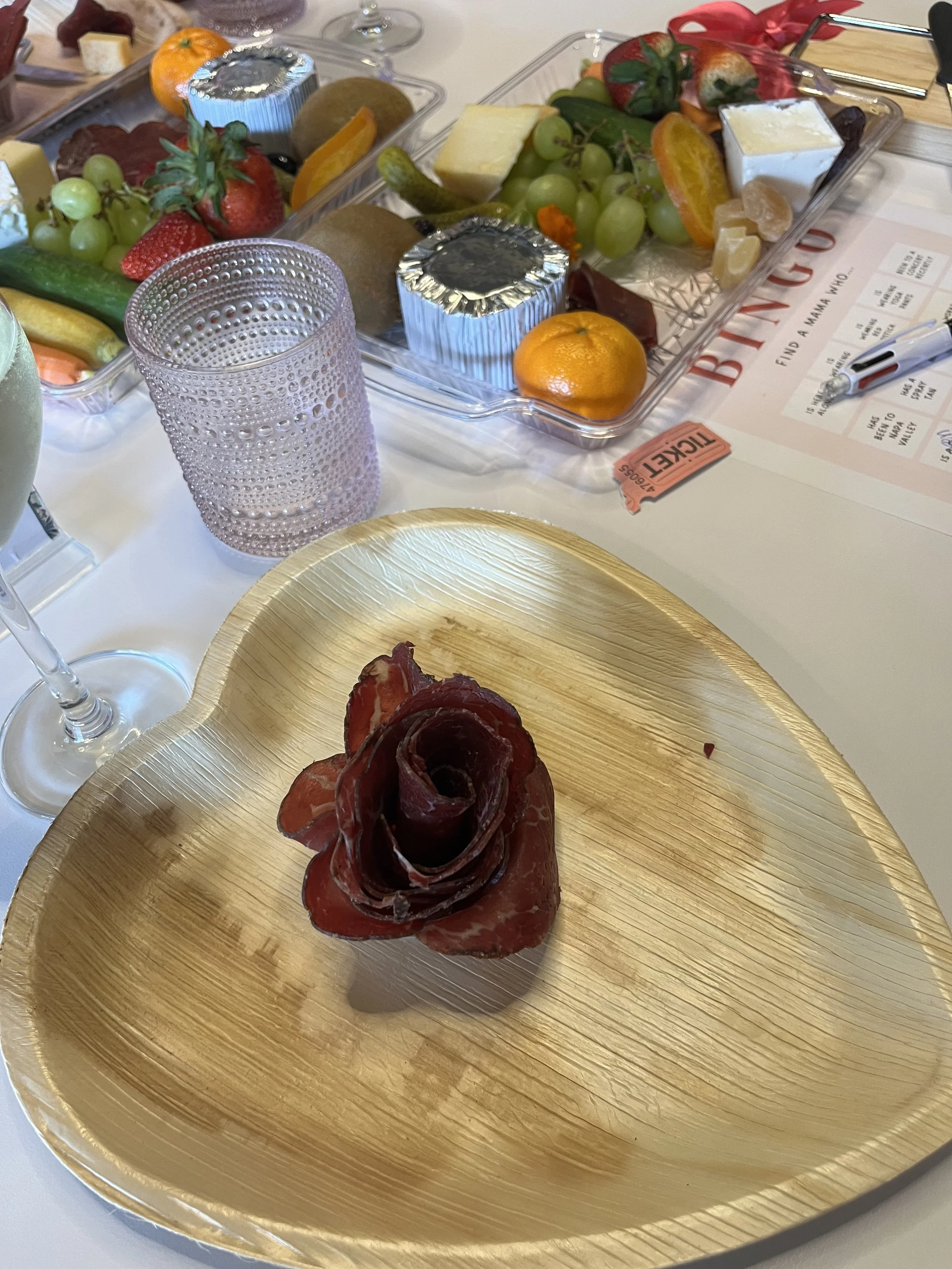 A gold heart-shaped plate with a rose-shaped arrangement of sliced cured meat, likely prosciutto, on it. In the background, there are trays of assorted fruits, cheeses, and wrapped items, along with a purple dotted glass and a pink ticket on a white 