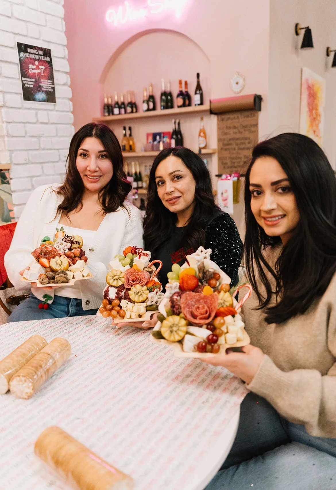 Three women celebrating Christmas, holding festive charcuterie boards with fruits and decorations, sitting at a decorated table in a cozy restaurant with wine bottles on a shelf in the background.