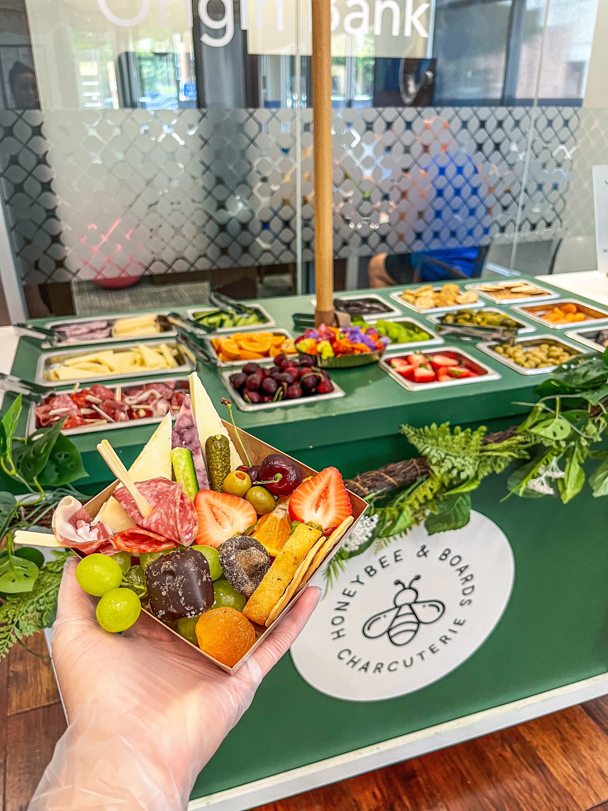 Person holding a small tray of assorted chocolates, strawberries, grapes, cheese, and crackers in front of a display of various sliced fruits and vegetables at a honey bee and boards charcuterie stand.