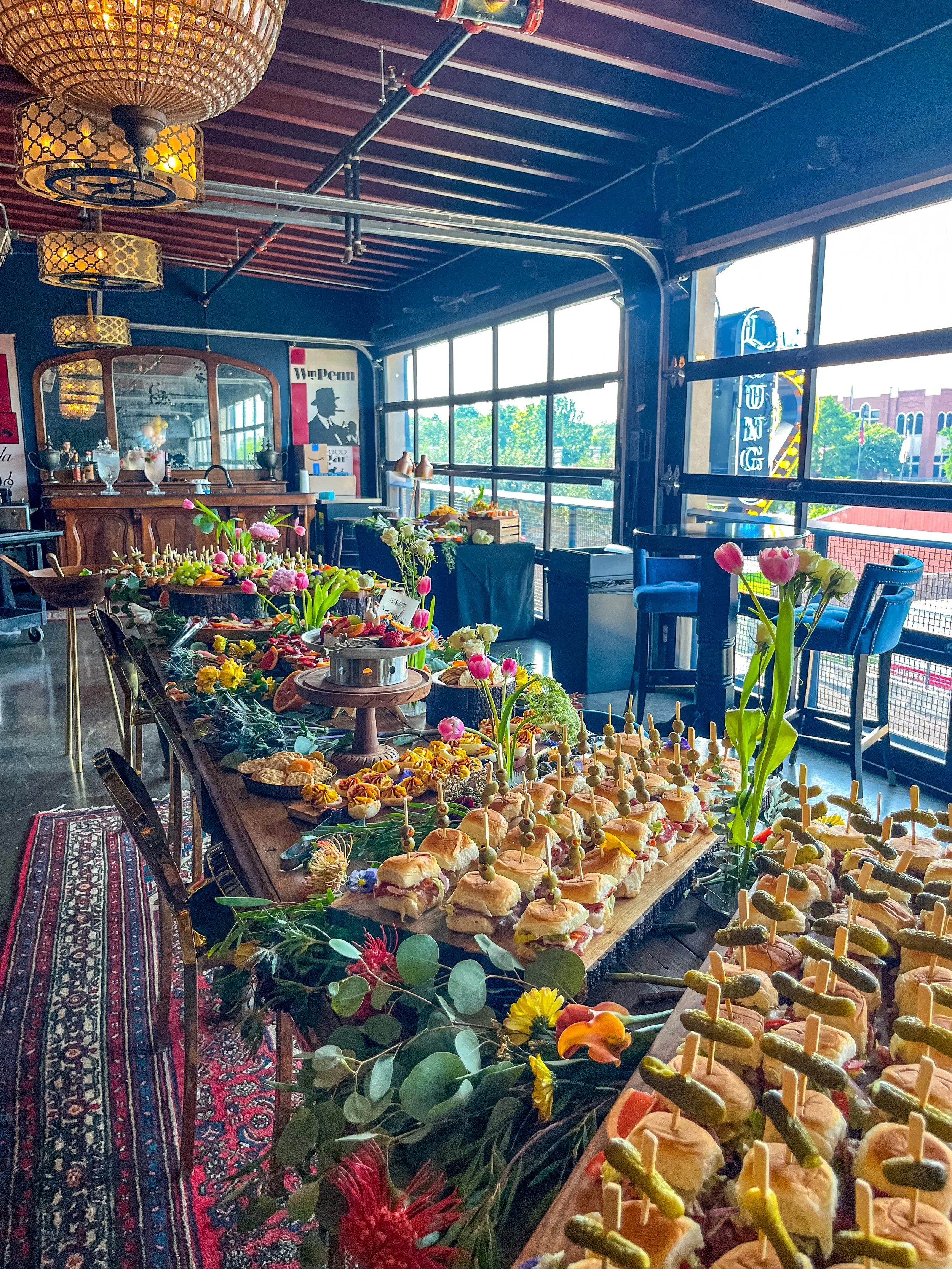 A decorated buffet table with assorted finger foods, sandwiches, and vegetable skewers, adorned with pink flowers and green foliage, in a room with large windows and vintage decor.