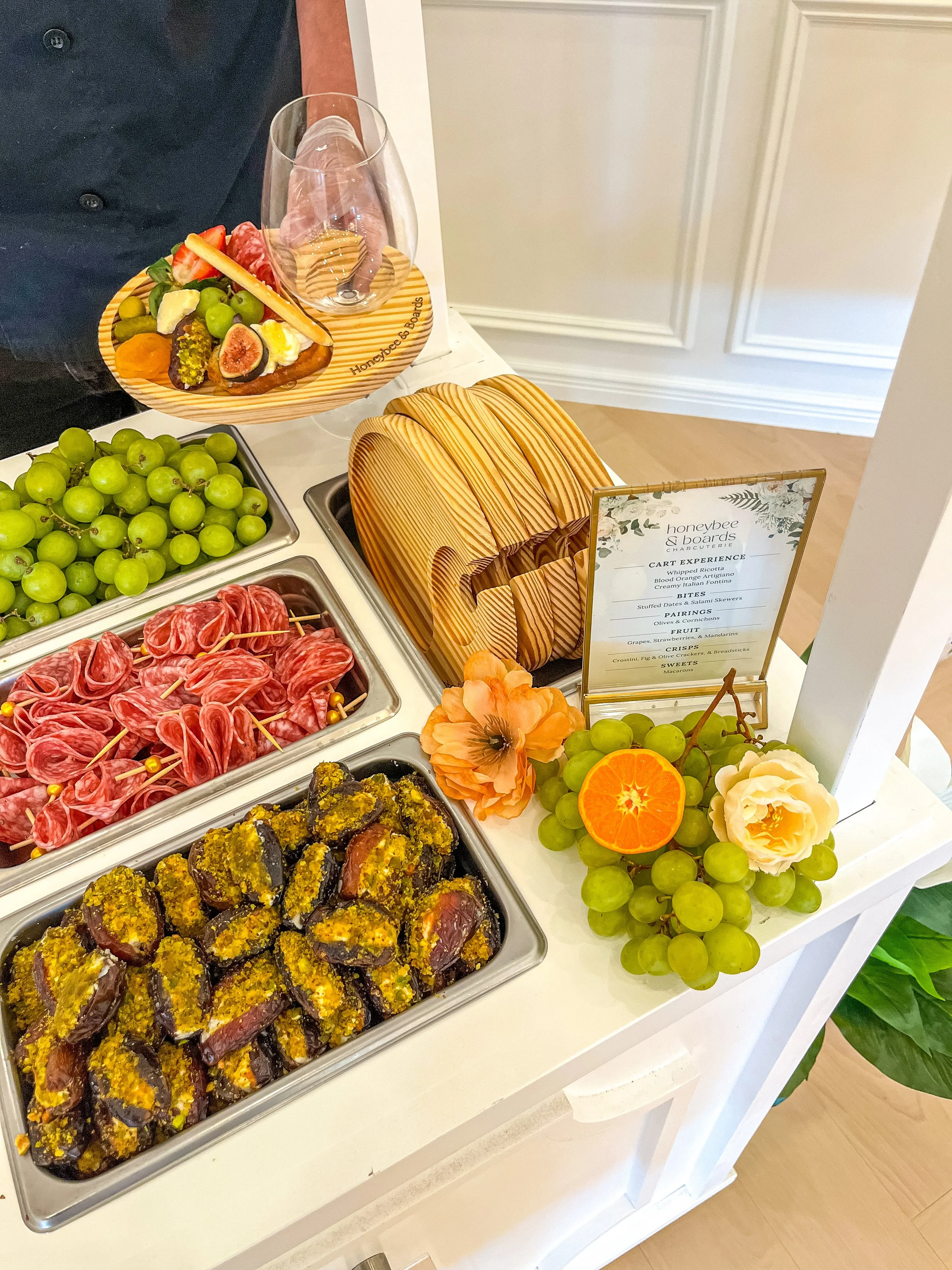A display of snacks including grapes, stuffed figs, and cured meats, along with a menu and decorative flowers at a buffet table.