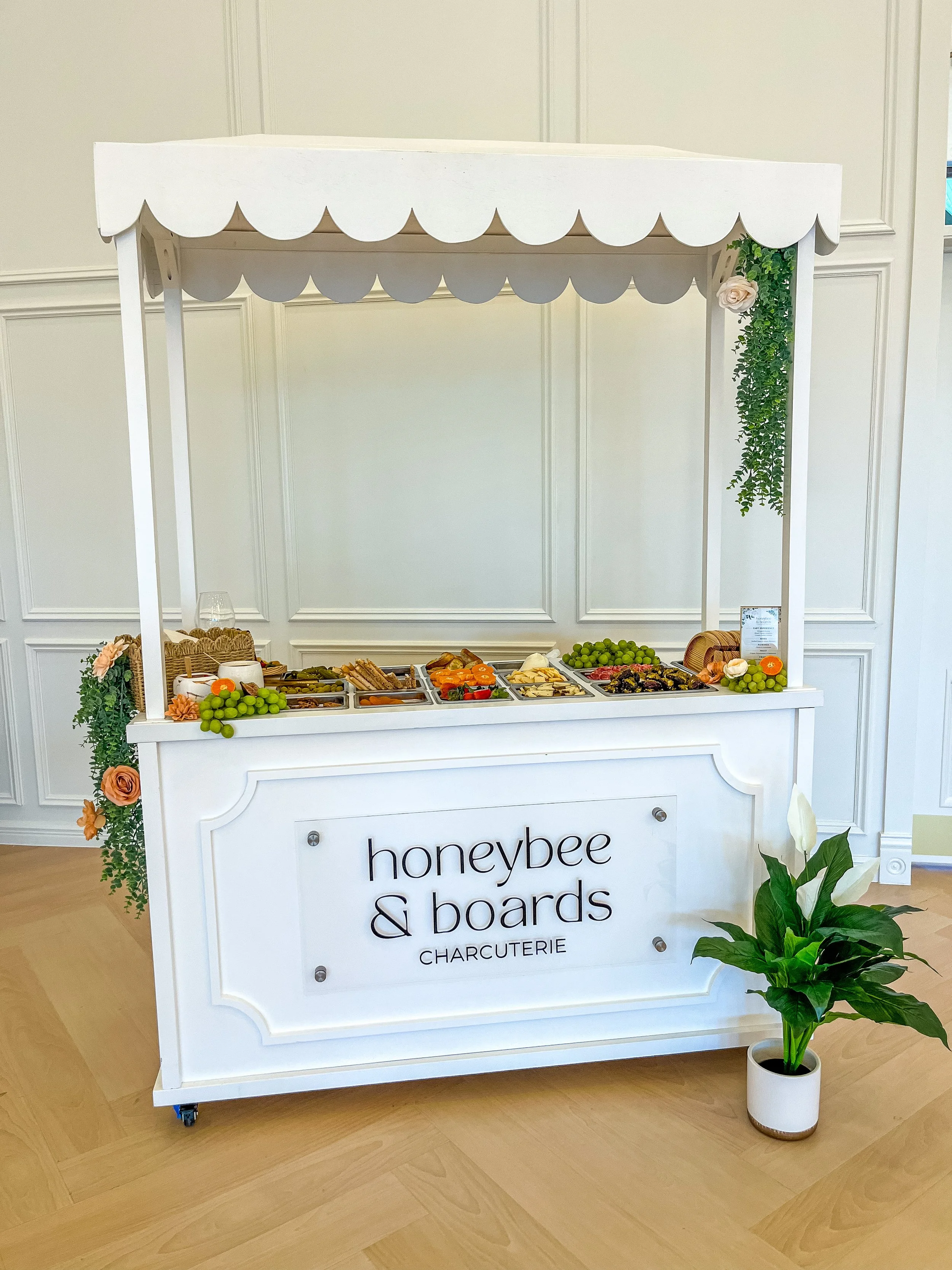A white charcuterie and cheese bar cart with the sign 'honeybee & boards' on the front, decorated with flowers and greenery, displaying various cheeses, meats, grapes, and crackers, with a small potted plant beside it.