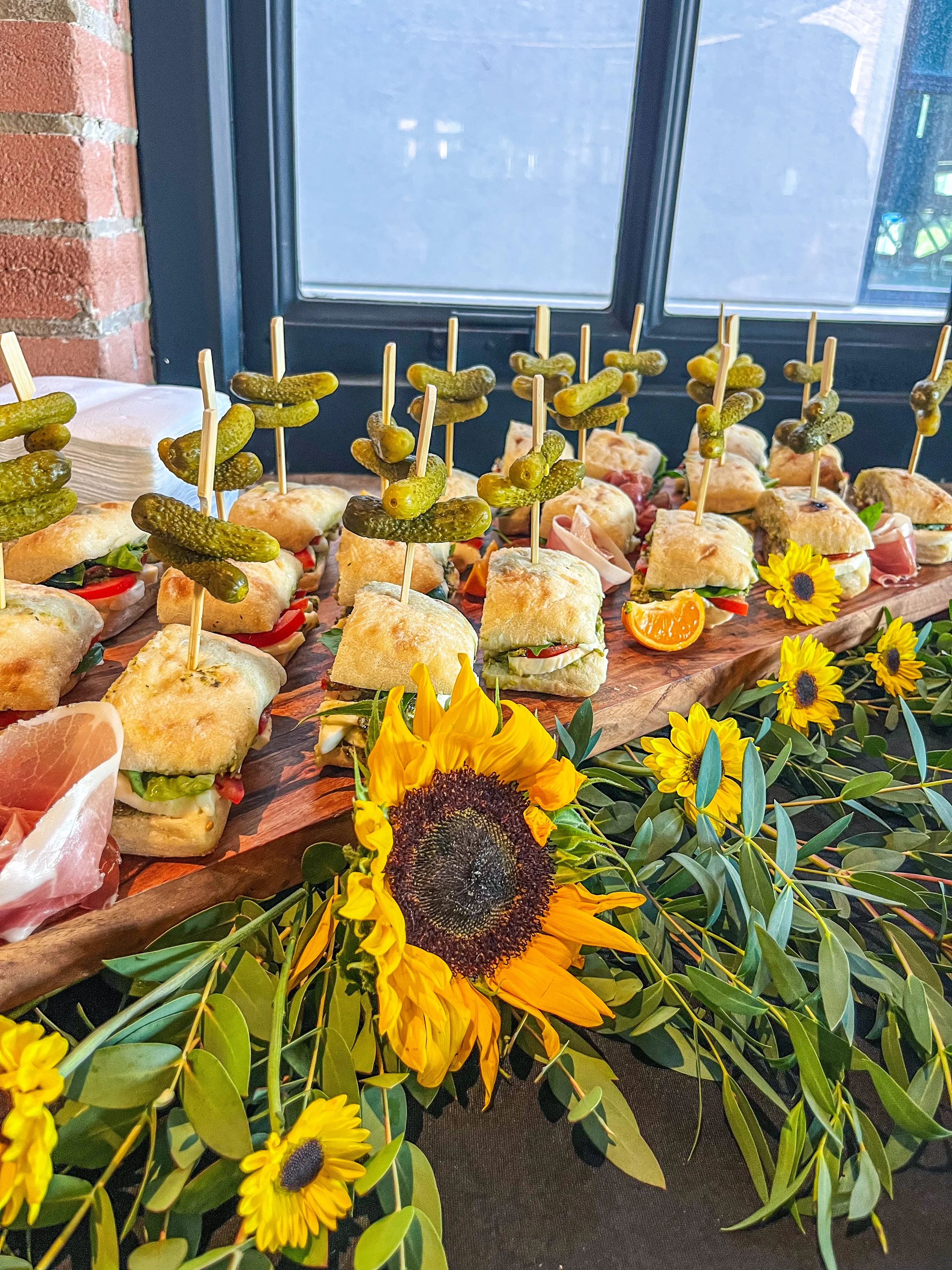 Assorted mini sandwiches with pickles on skewers, garnished with sunflowers and yellow flowers on a wooden tray.