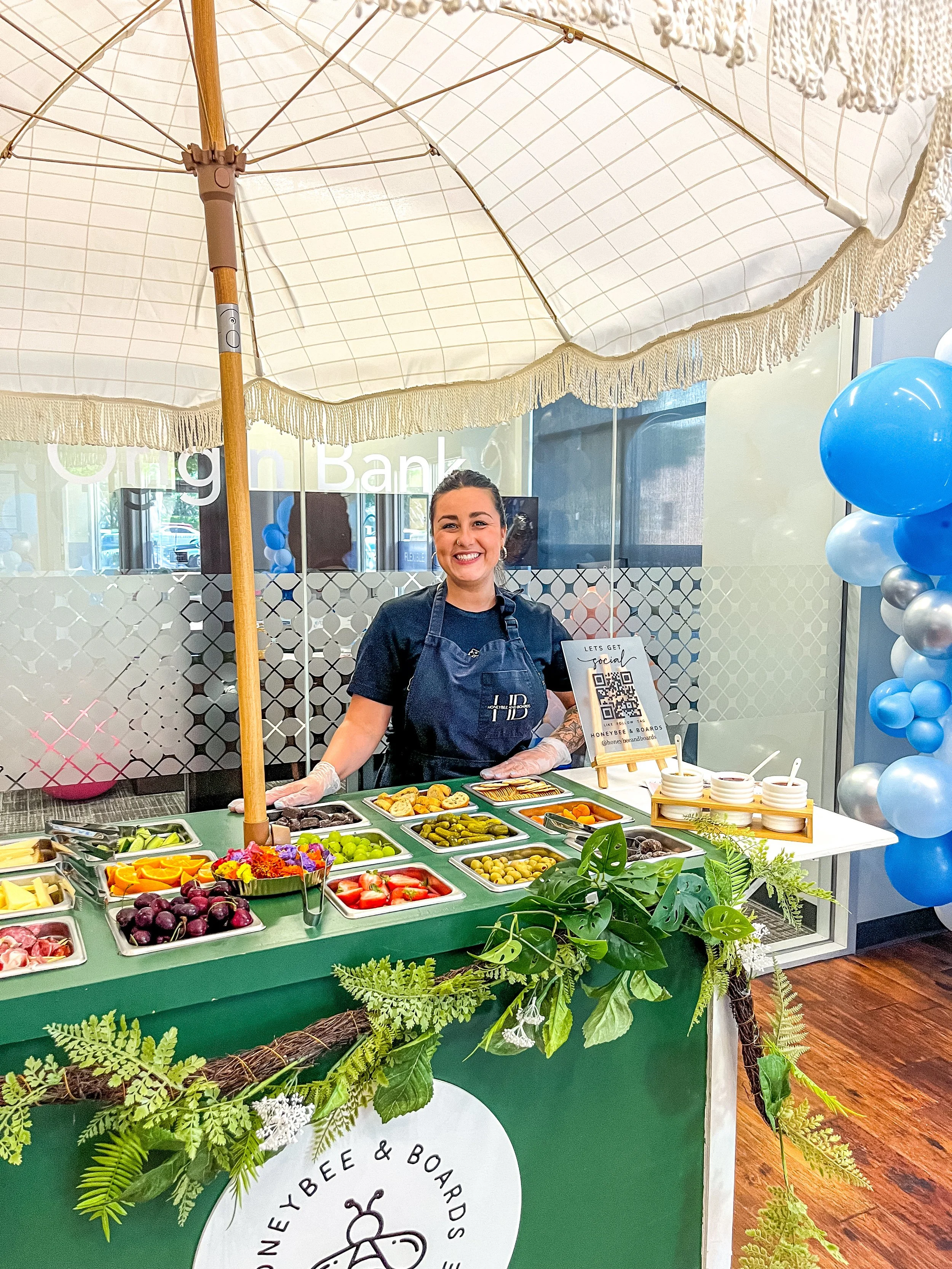 A smiling woman behind a colorful fruit and snack stand decorated with greenery, under a large white umbrella, with balloons on the side, at an indoor event or market.