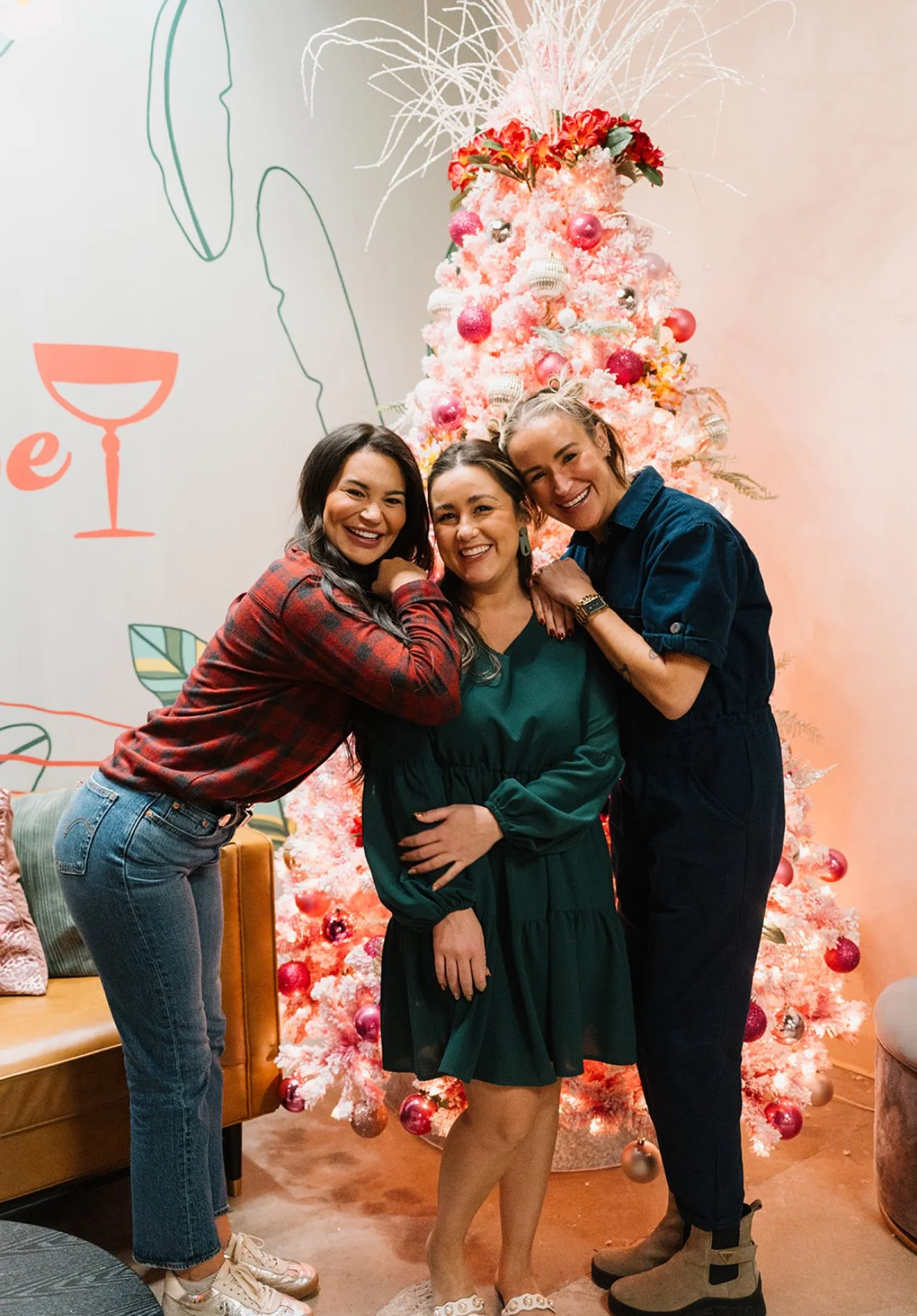 Three women smiling and hugging in front of a decorated pink Christmas tree with ornaments, in a festive indoor setting.