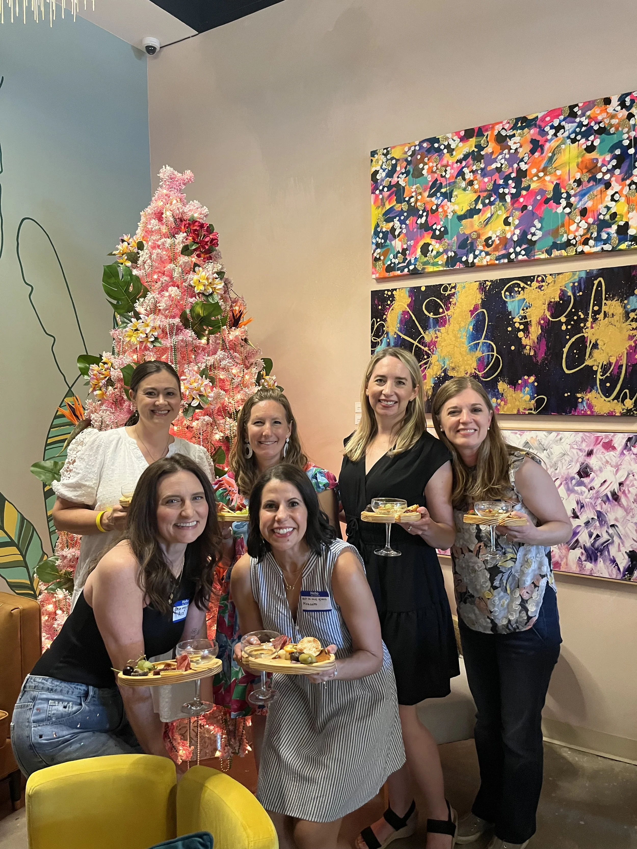 Group of six women celebrating, holding dessert glasses with cheese platter, in a room decorated with colorful abstract paintings and a pink Christmas tree, at an art gallery or modern event space.