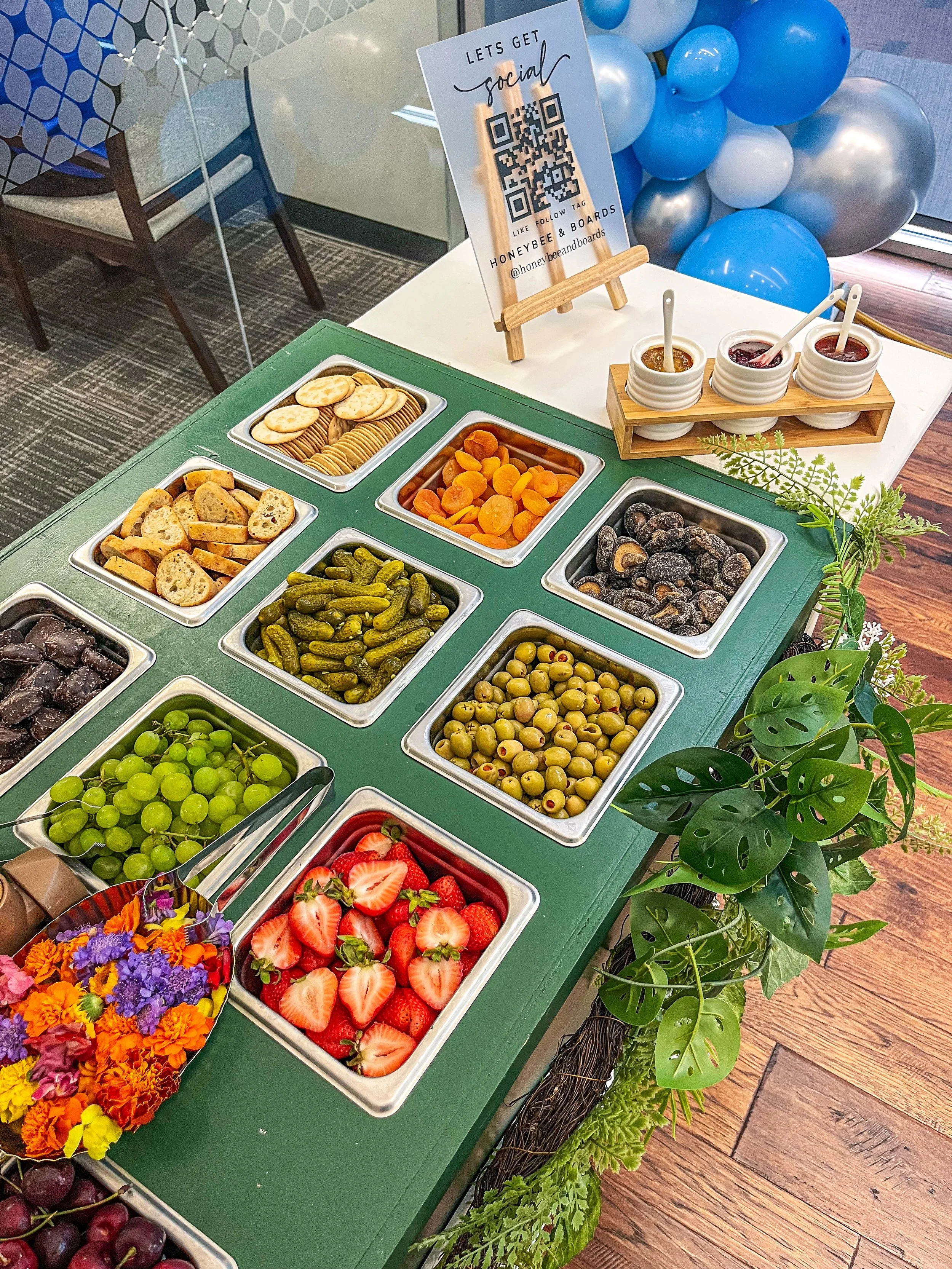 A dessert and snack table with bowls of strawberries, green grapes, black grapes, pickles, dried apricots, various types of cookies, and chocolates. There is a sign with a QR code for social media and a tray of three small bowls with different toppin