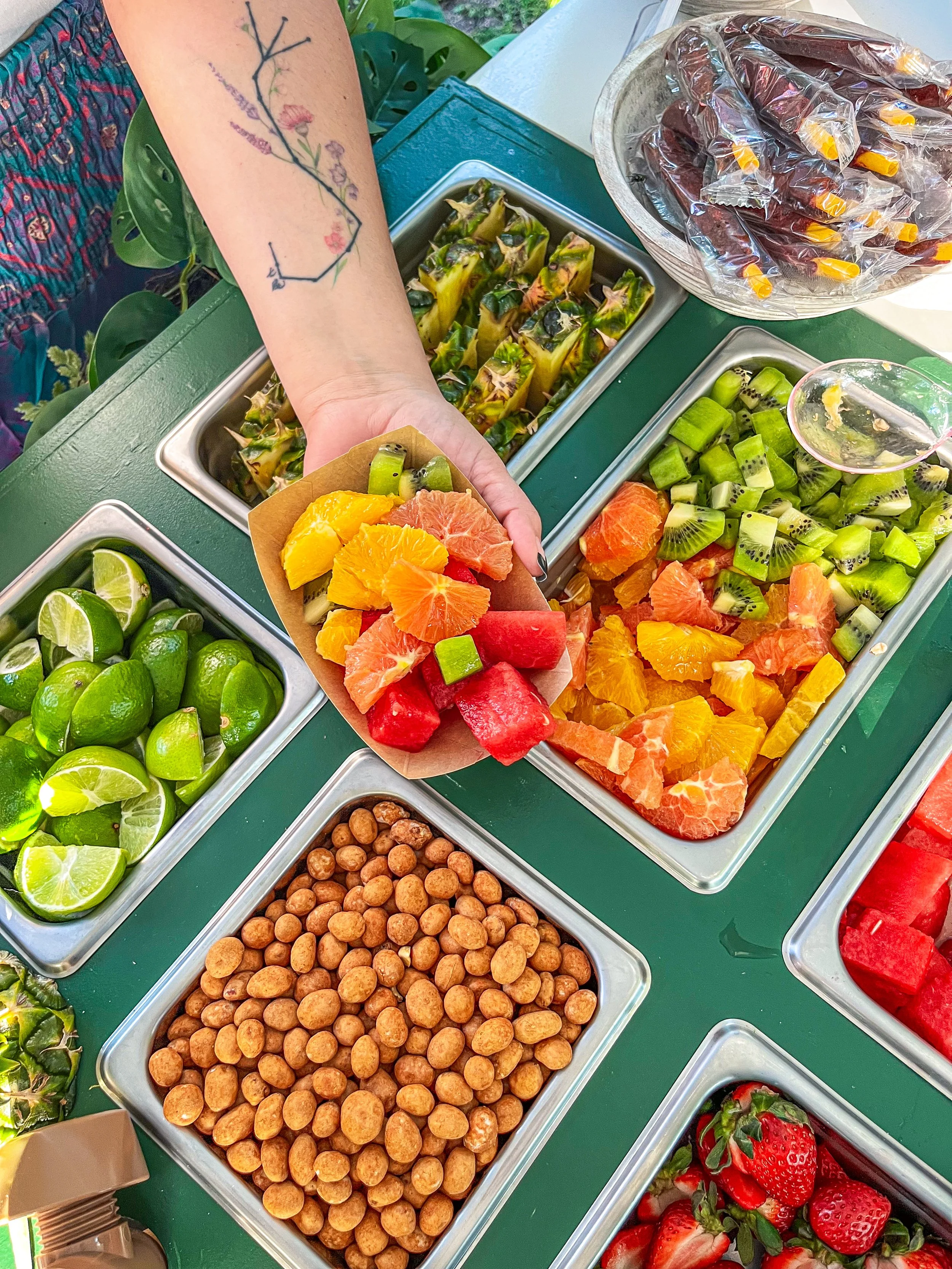 A person scooping cut-up assorted fruits such as watermelon, orange, grapefruit, and honeydew melon from a buffet-style fruit bar. The table also has containers of strawberries, kiwifruit, lime wedges, and chickpeas. The person's arm has a floral tat