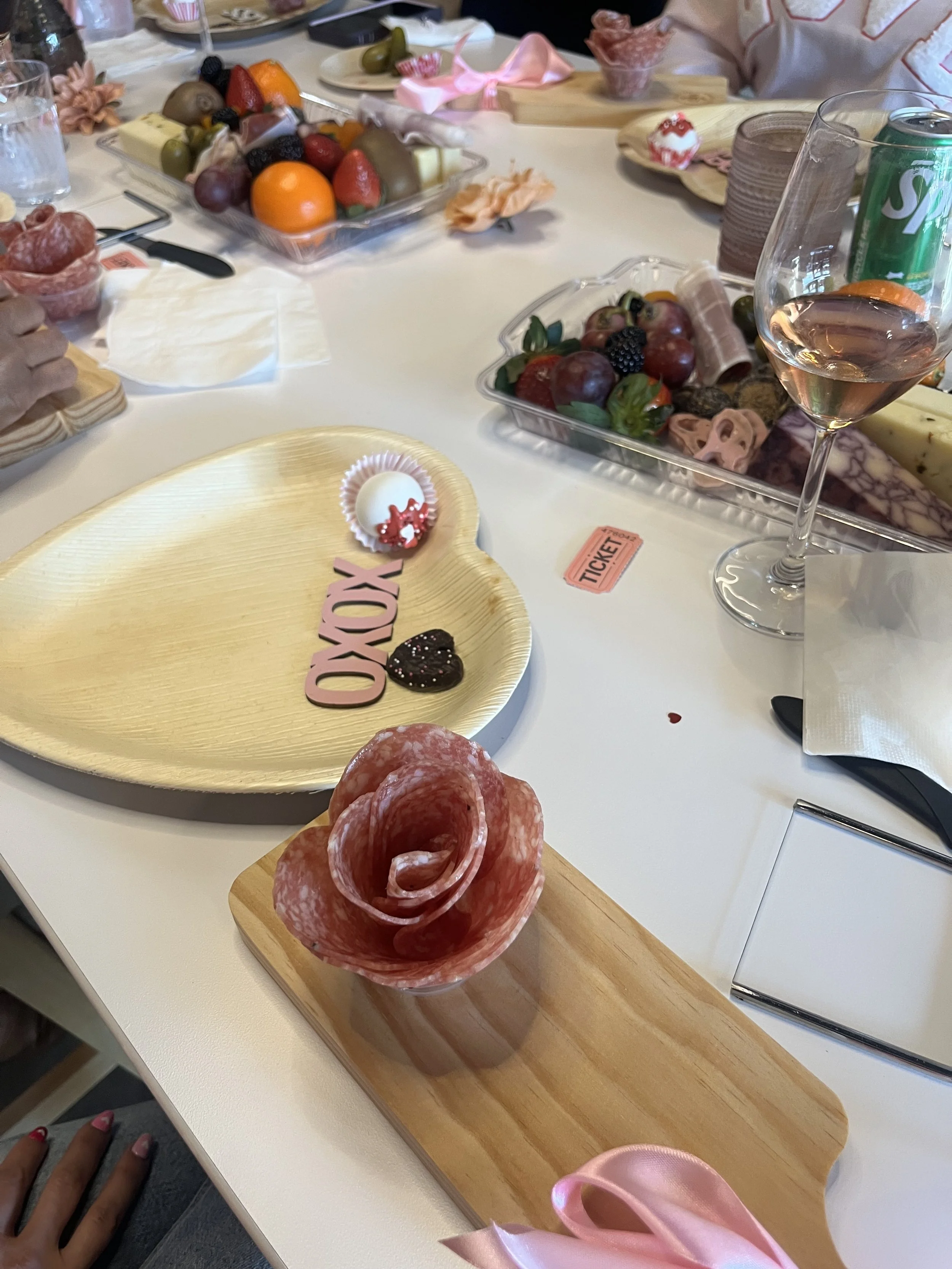 A table set with assorted fruits, salami slices shaped like a rose, chocolates, a glass of rosé wine, a can of Sprite, and a center arrangement of fruit and chocolates. There are decorative pink ribbons and a 'XOXO' sign on a small dish.