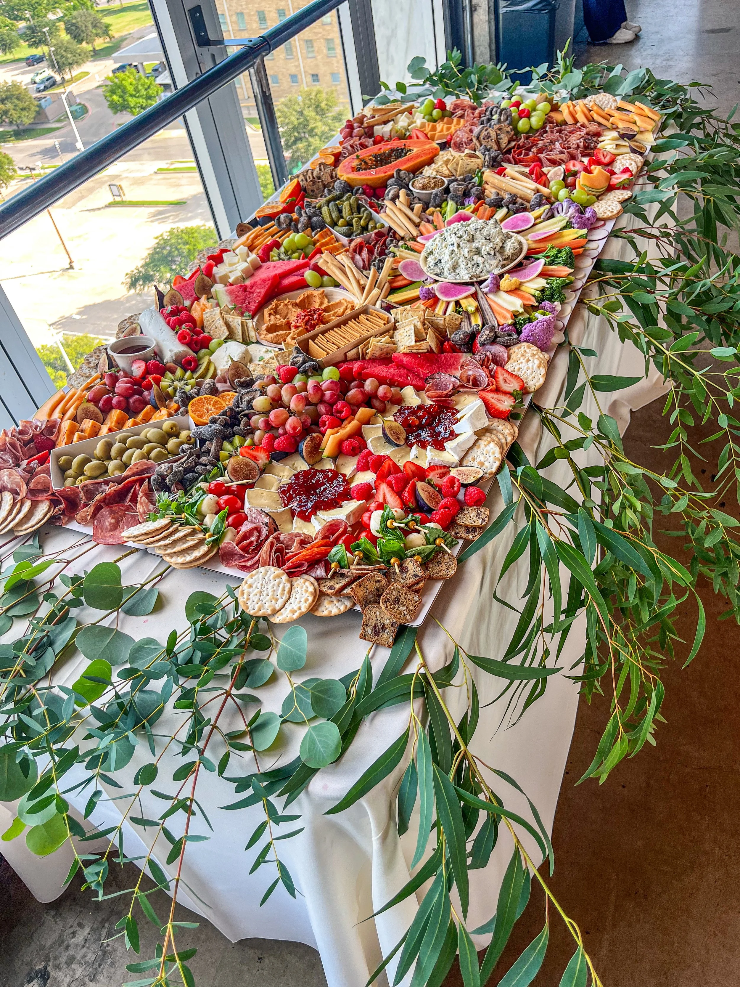 A long table decorated with greenery and laden with assorted cheeses, crackers, fruits, vegetables, meats, and dips, set up for a buffet or party celebration in a high-rise venue with large windows overlooking a cityscape.