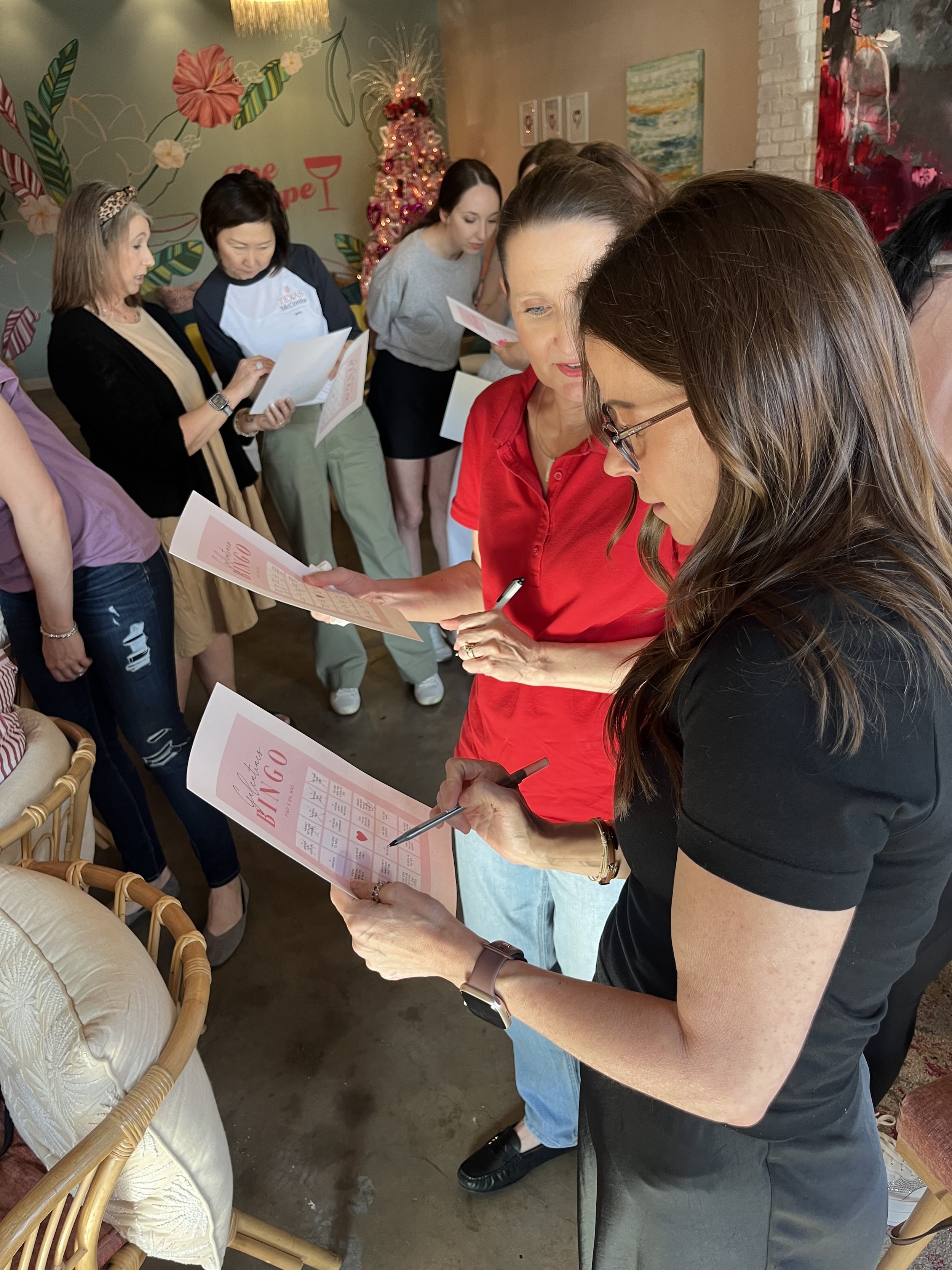 A group of women participating in a bingo game indoors, some are reading their bingo cards, one woman is writing, and others are holding their cards. There is a decorated Christmas tree and wall art in the background.