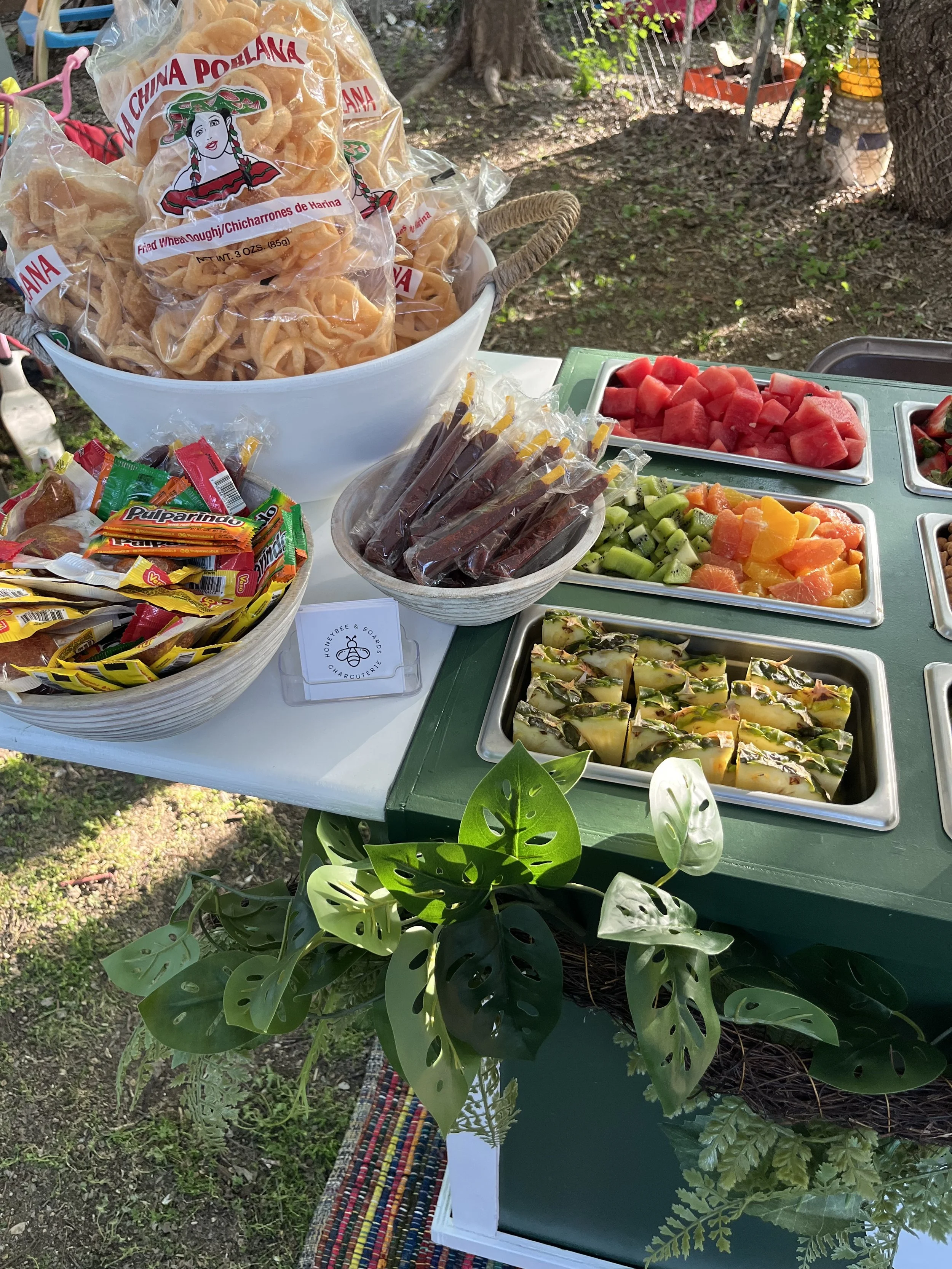 Assorted fresh fruits including watermelon, kiwi, mango, papaya, and sliced vegetables displayed on a table outside. There are packaged snacks, a bowl of pink-colored snacks, and a green leafy plant for decoration.