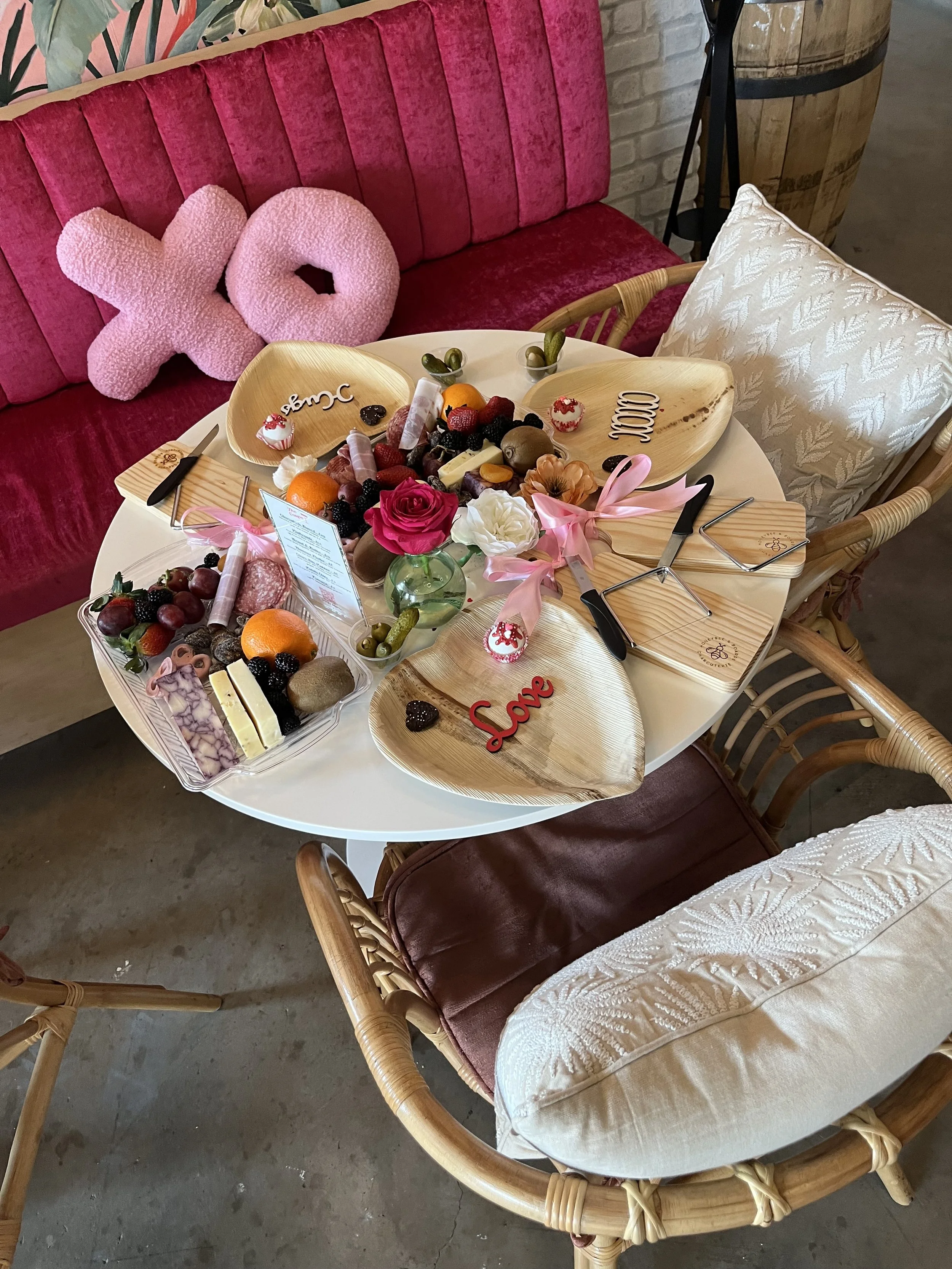 A table set with various desserts, a small bouquet of flowers, and decorative wooden plates with words like 'Love' and 'Sweet'; surrounded by a pink velvet sofa with large pink plush letters 'XOXO' and cozy cushions.
