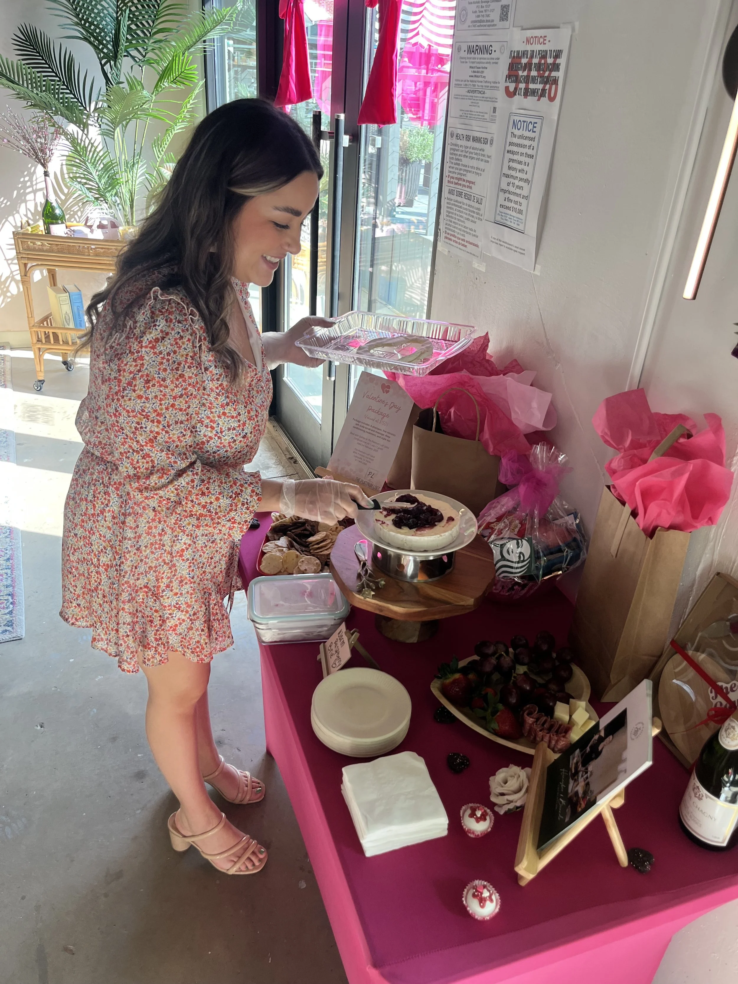 Young woman in a floral dress serving herself dessert at Valentine's Day event decorated with pink tissue paper and treats on a table.