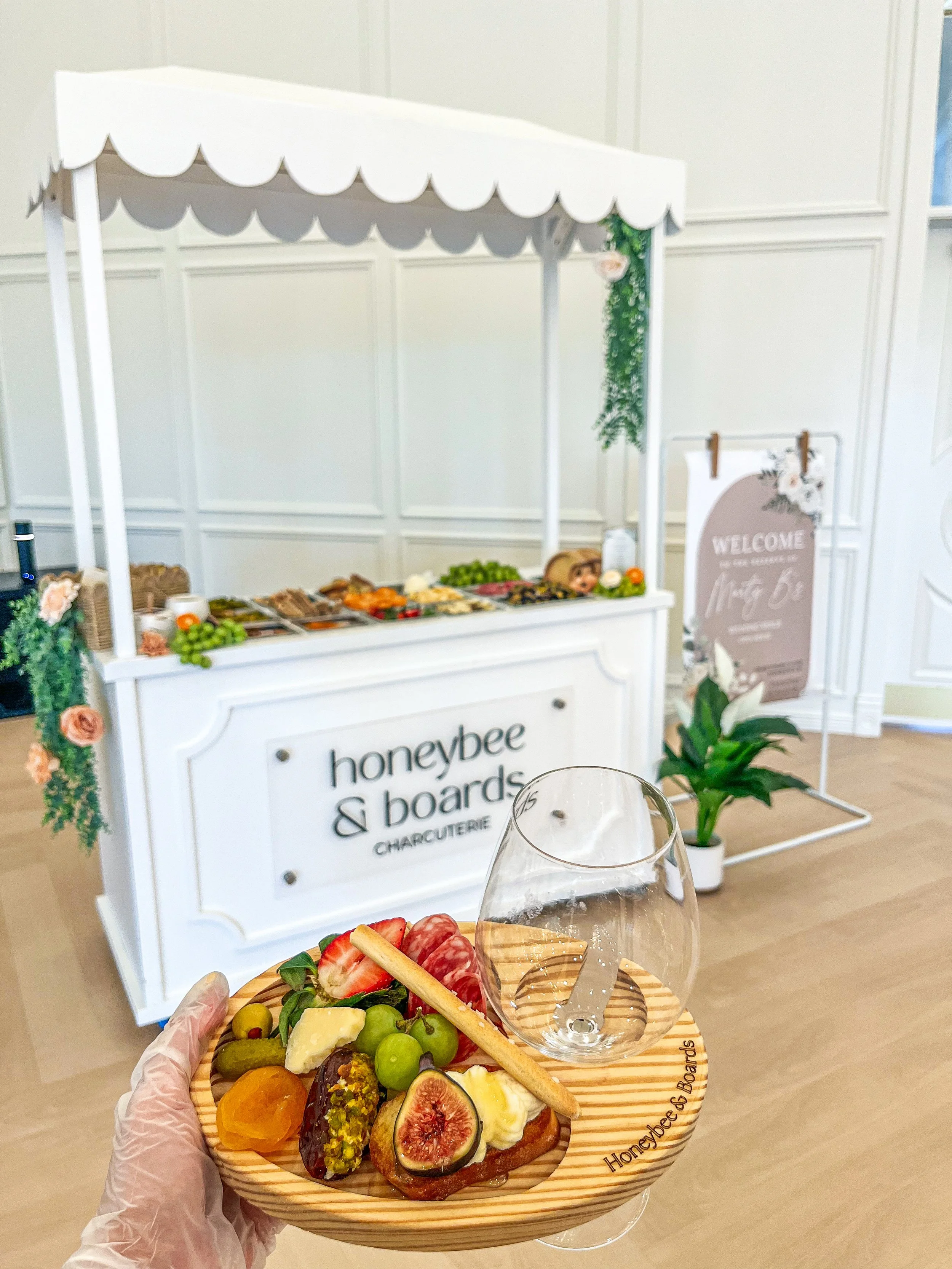 A person holding a wooden tray with assorted fruits, a biscuit, and a glass of white wine in front of a white charcuterie stand decorated with flowers. The stand has the sign 'honeybee & boards'.