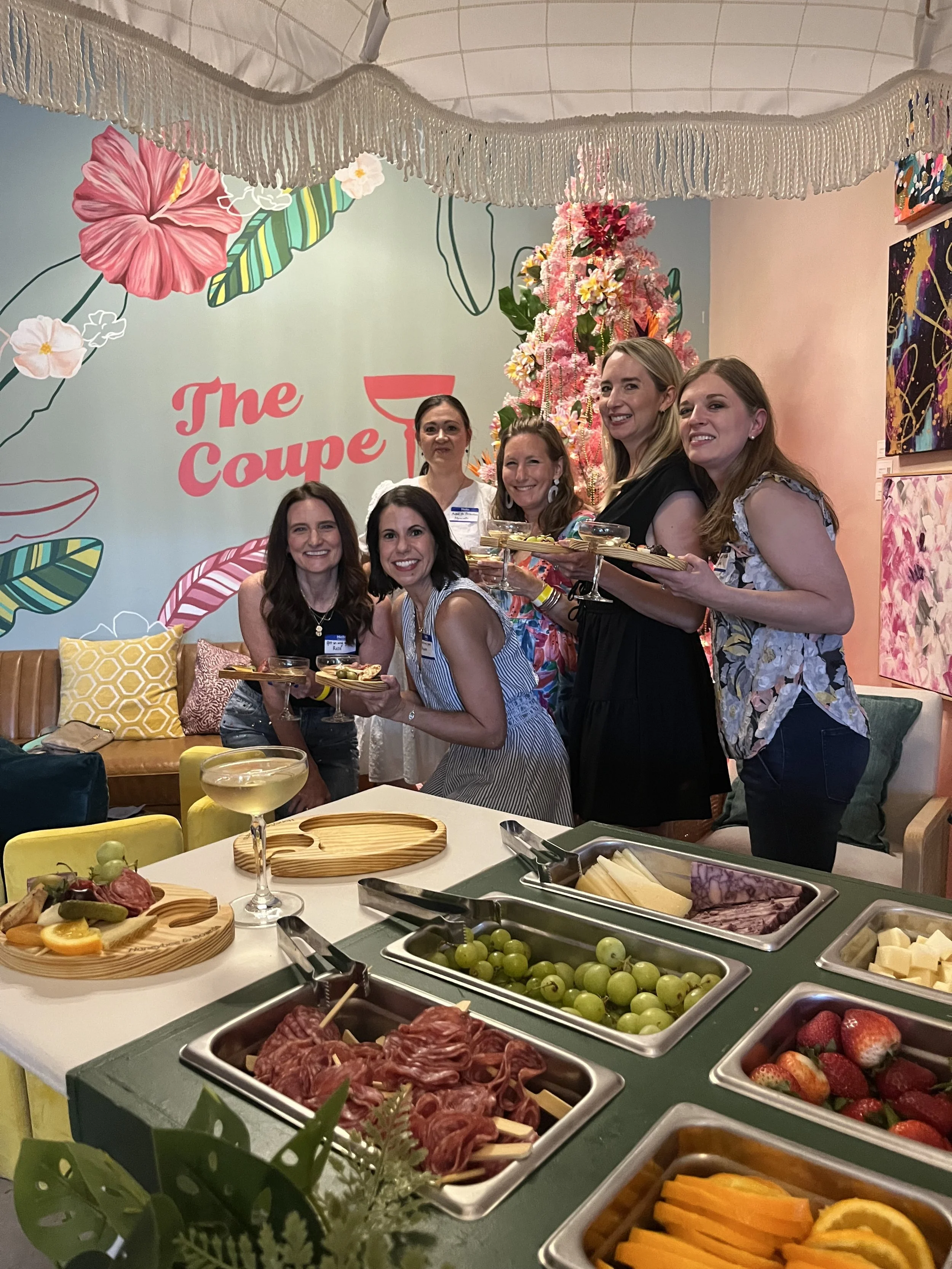 Six women gathering at a cocktail party, holding plates of food, standing in a decorated room with colorful wall art and flowers, with a table of cheese, fruits, and charcuterie in the foreground.