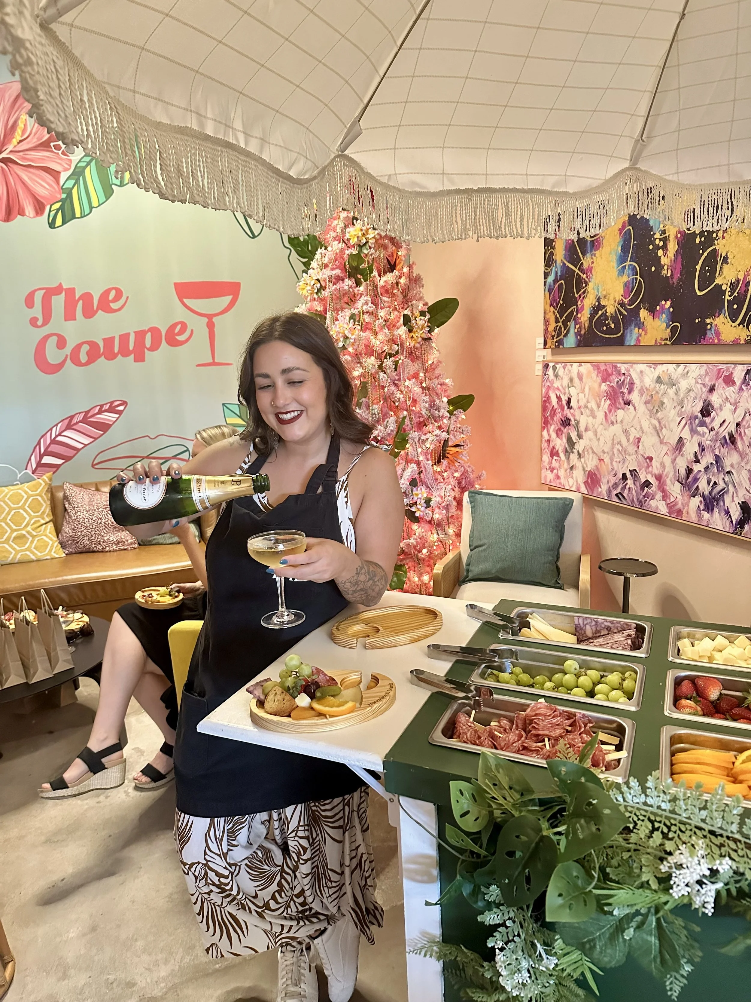 A woman in a black apron pours champagne into a glass at a festive gathering with a colorful floral and abstract art decor, a Christmas tree, and a buffet of fruits and snacks.