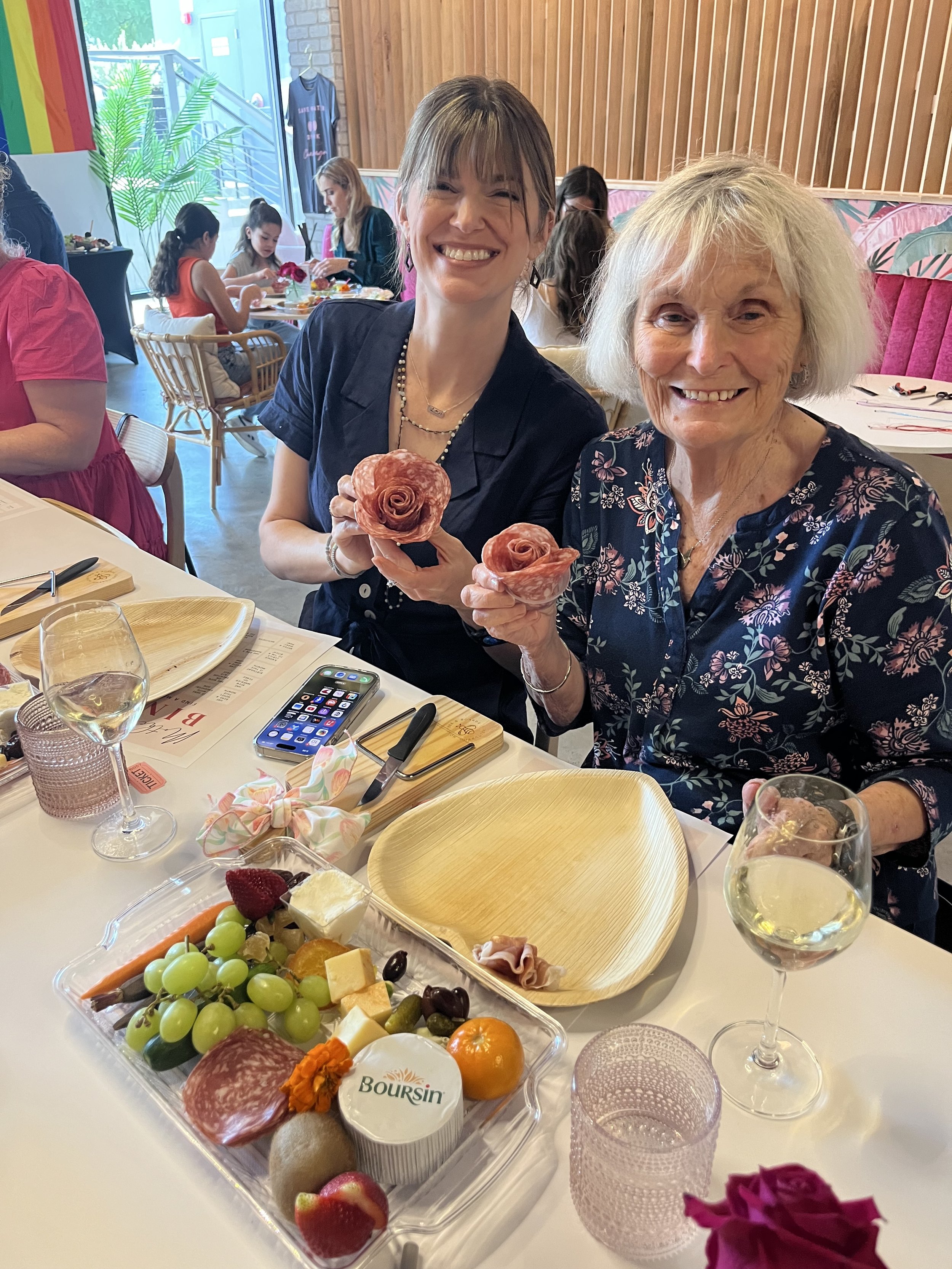 Two women sitting at a table with a charcuterie platter, holding rose-shaped meats, smiling in a bright restaurant with other diners in the background.