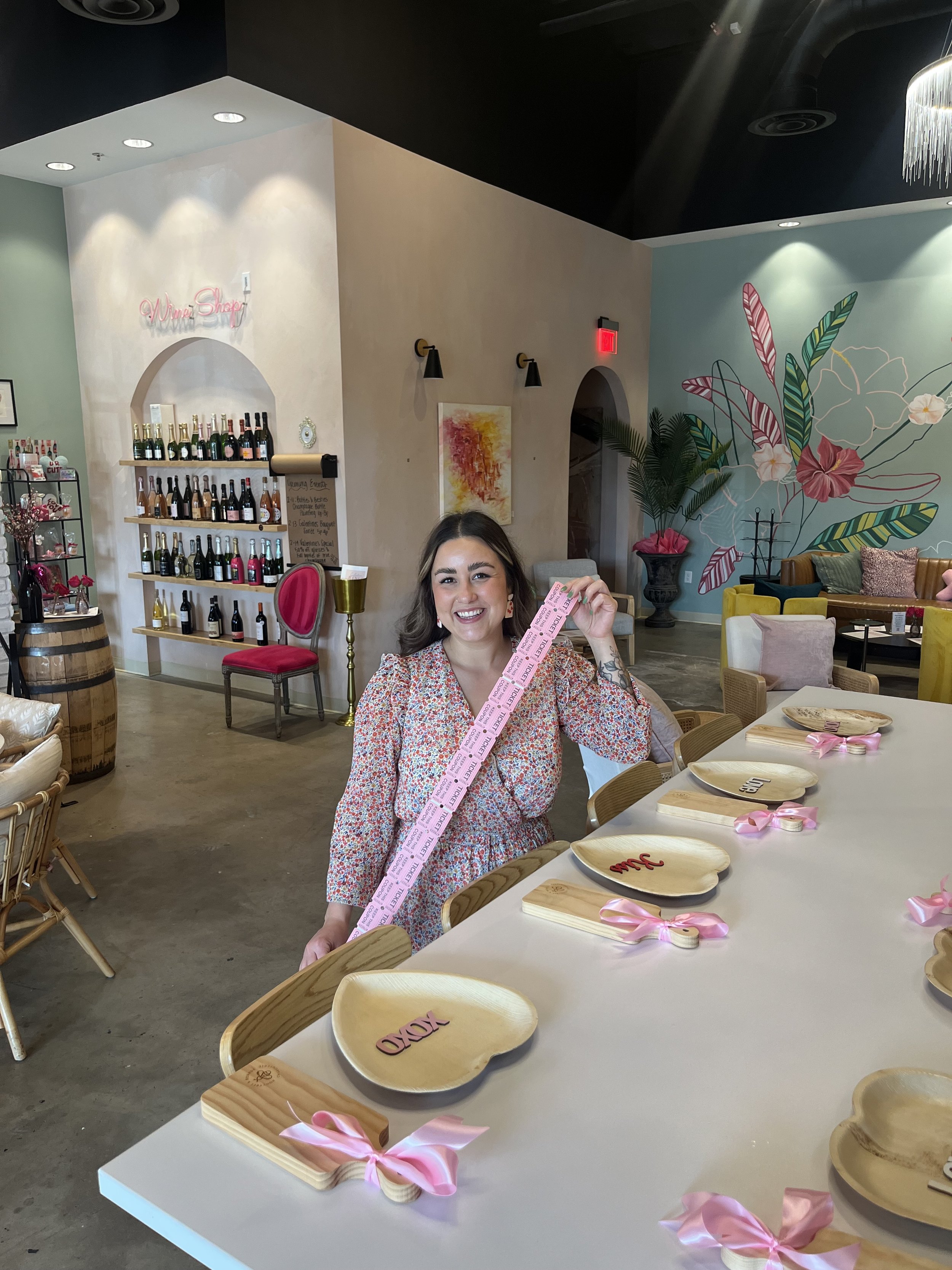 A woman smiling and holding a pink ticket and a pink ribbon, sitting at a long table decorated with wooden platters with names on them and pink ribbons tied to small wooden handles, inside a colorful, floral-themed space with wine bottles on shelves 