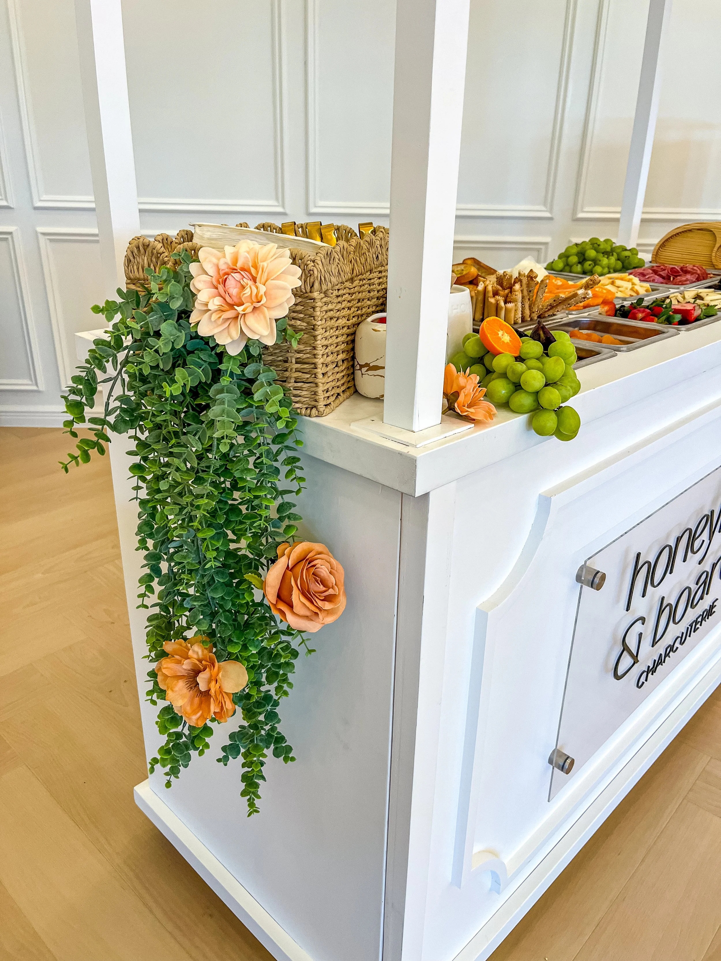 A white food cart decorated with a cascading arrangement of greenery and peach-colored flowers, with tray of fresh fruits and snacks on top.