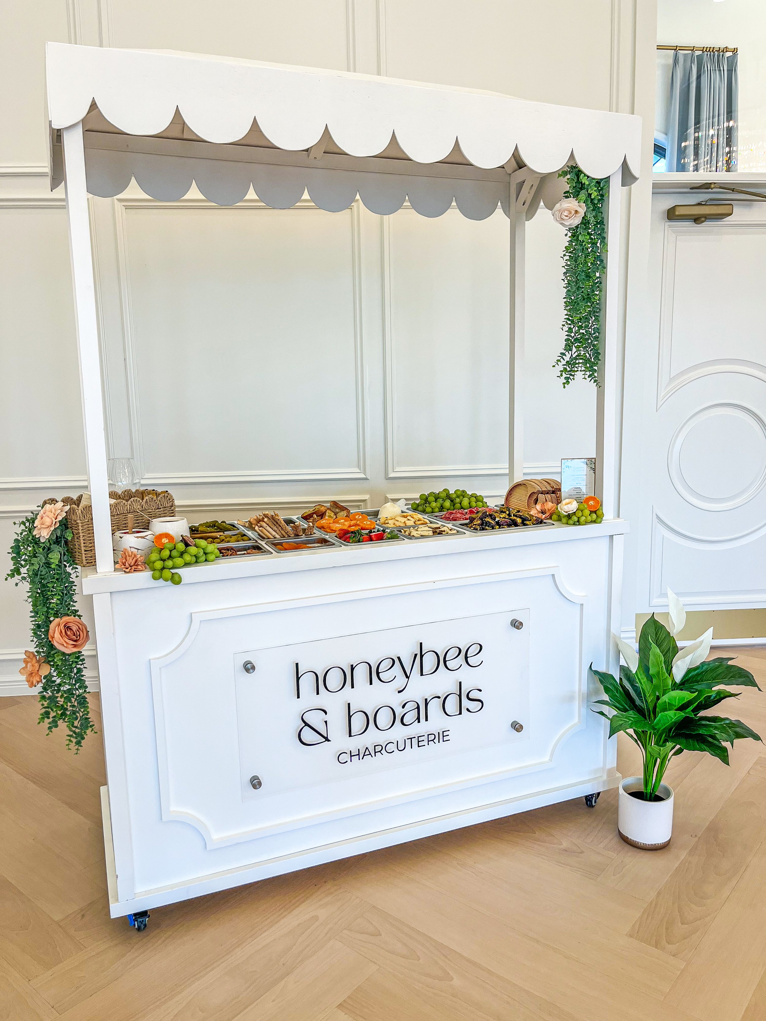 A white charcuterie stand with a canopy, decorated with flowers and greenery, featuring an array of food items such as cheeses, grapes, meats, and crackers, with a potted plant beside it.