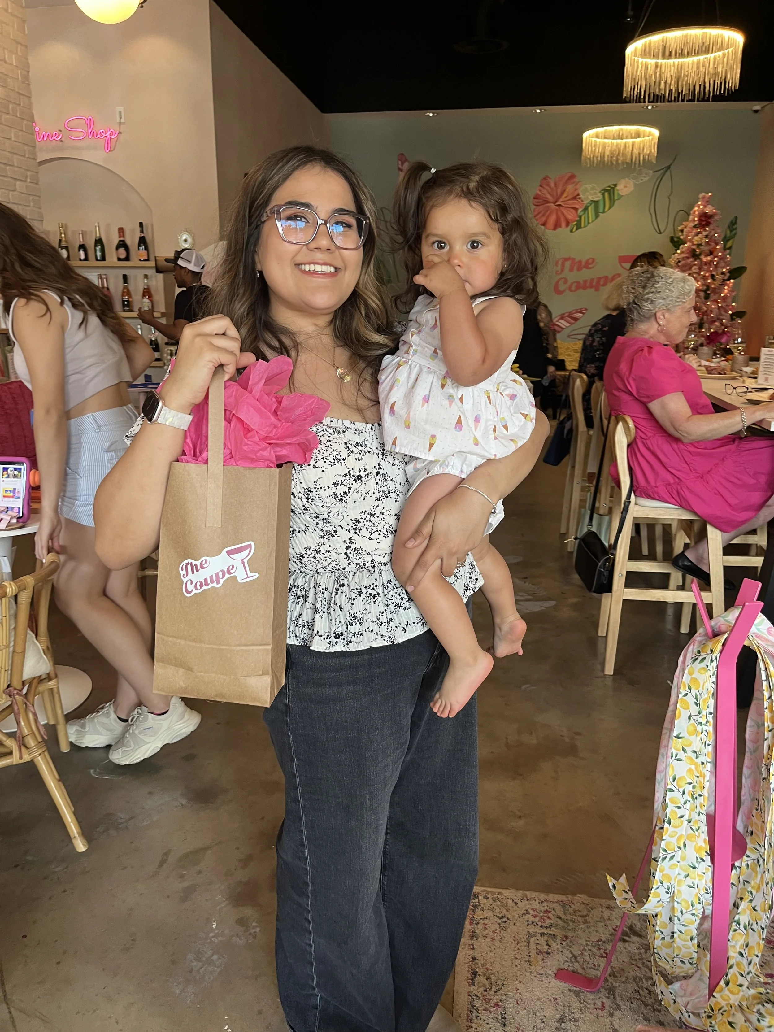 A woman smiling and holding a young girl in an indoor setting with decorated walls and people seated at tables. The woman carries a gift bag with pink tissue paper.