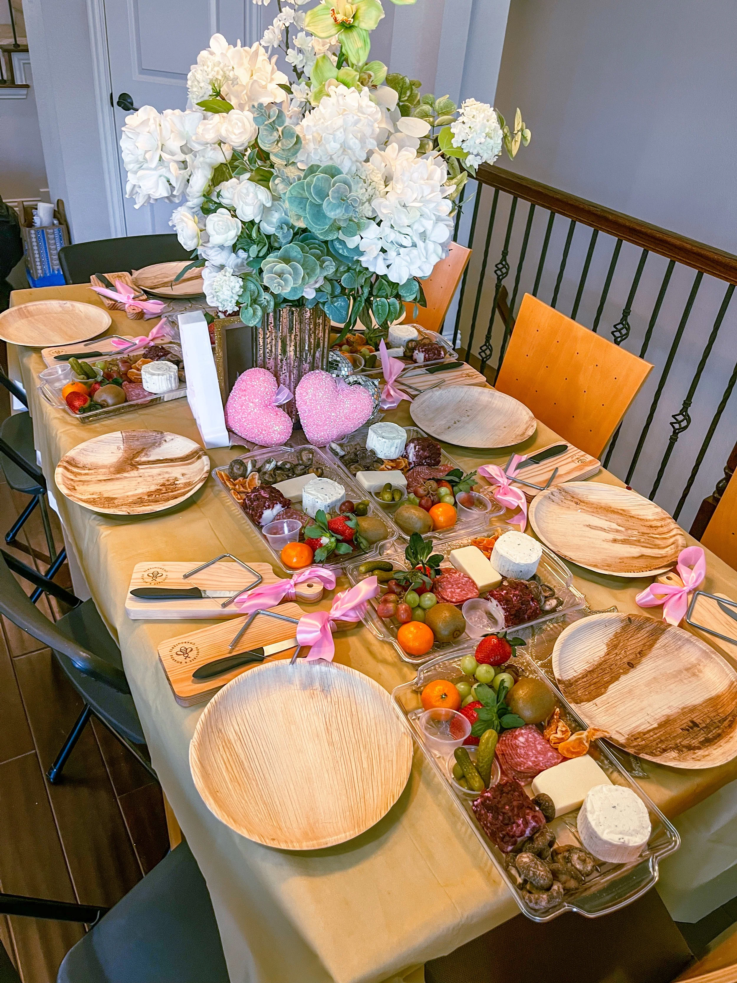 A table set for a celebration with a large floral centerpiece of white and pale green flowers, surrounded by wooden plates, cheese, various fruits, meats, and pink ribbon decorations.