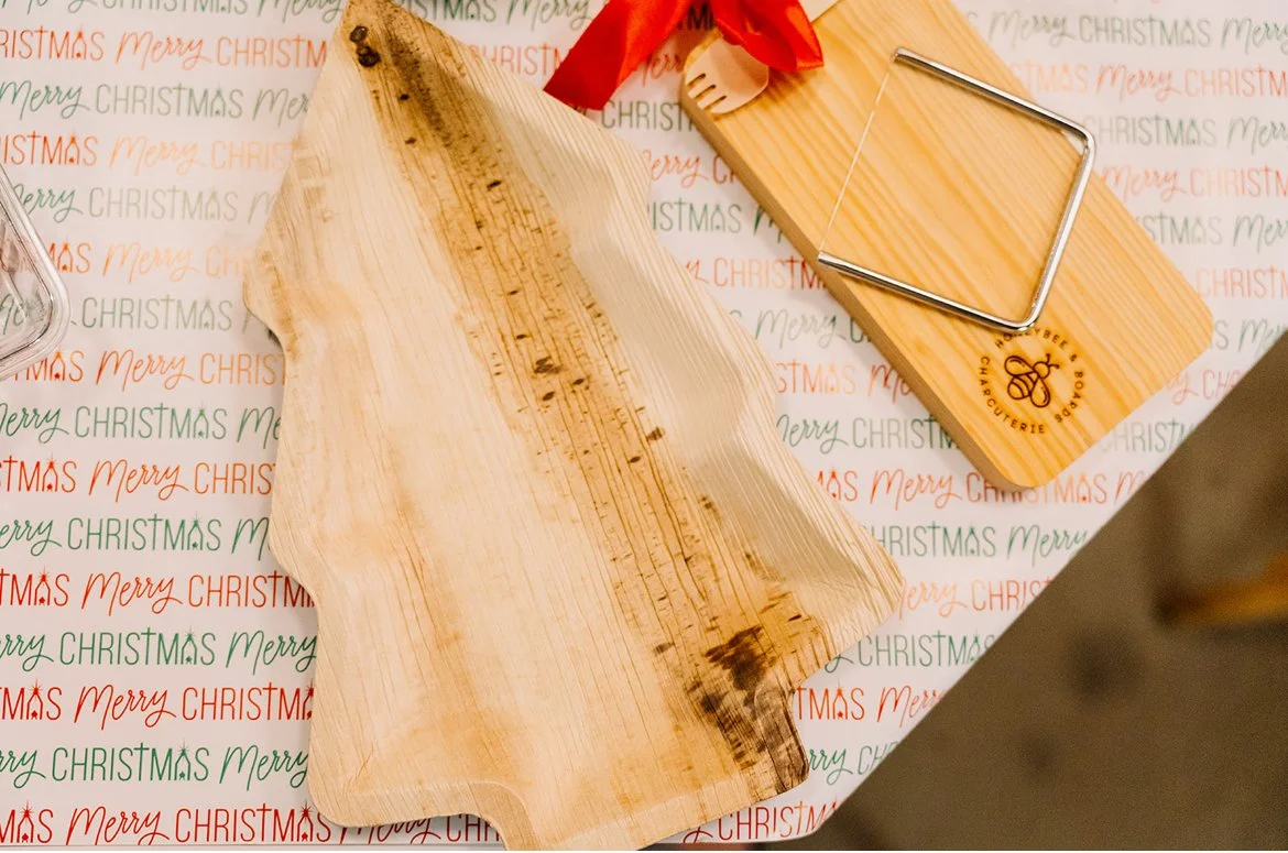 A piece of wooden cutting board with a natural edge, next to a smaller wooden cutting board with a corkscrew logo, on festive Christmas wrapping paper.
